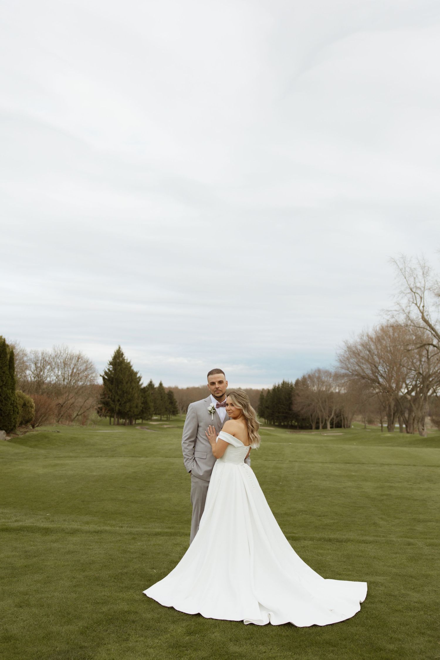 Bride and groom pose on a lush green golf course, her white gown trailing elegantly behind them.
