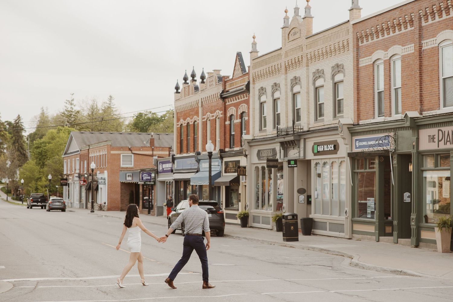 Couple holding hands crossing a charming small-town street lined with historic brick storefronts.