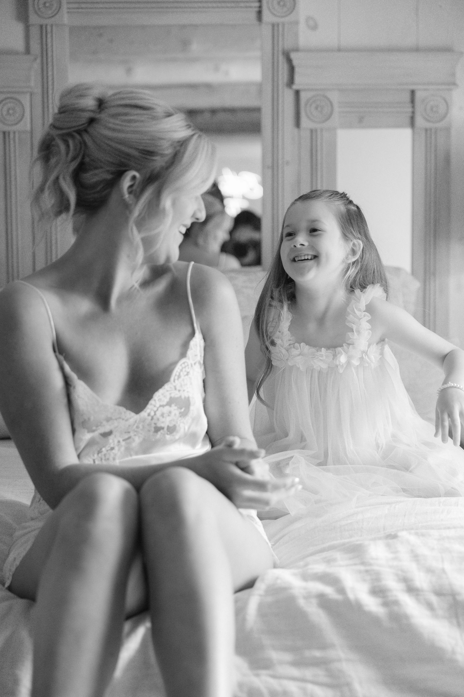 Black and white photo of a bride in lace sitting on a bed smiling with a young flower girl in a white dress.