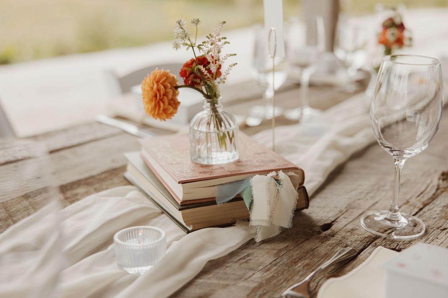 Rustic wedding table centerpiece featuring stacked books, a bud vase with colorful flowers, and glassware.