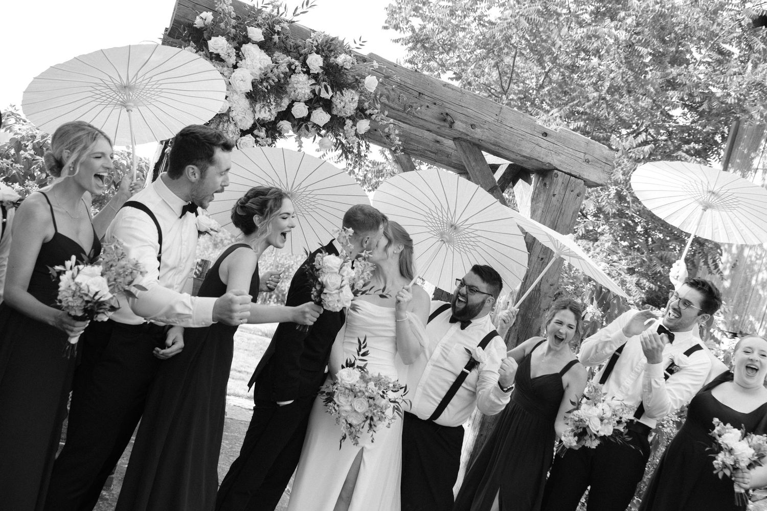 Black and white wedding party photo with bridesmaids and groomsmen holding white parasols outdoors.