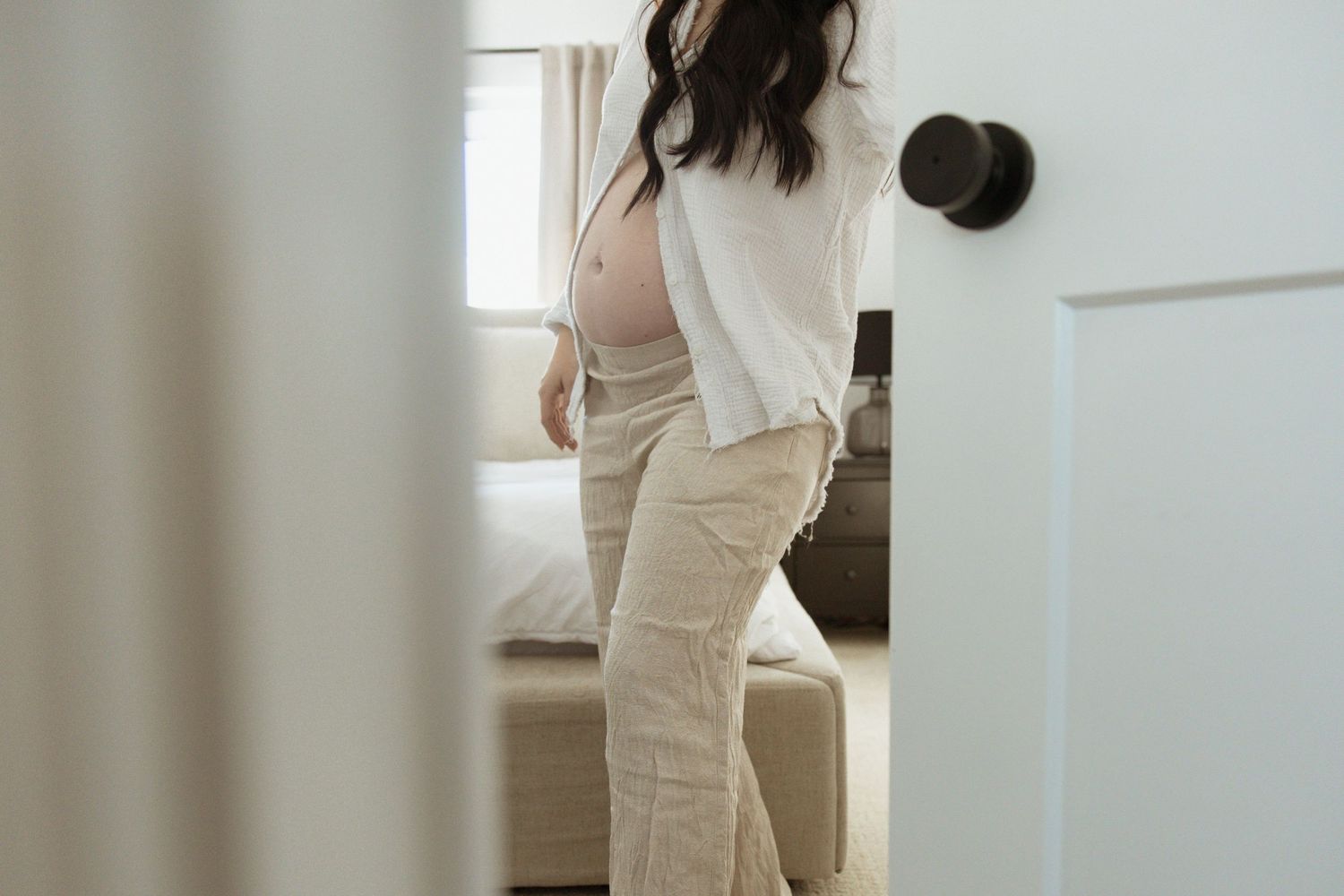 Pregnant woman in a white shirt and beige pants standing in a softly lit bedroom doorway showing her baby bump.