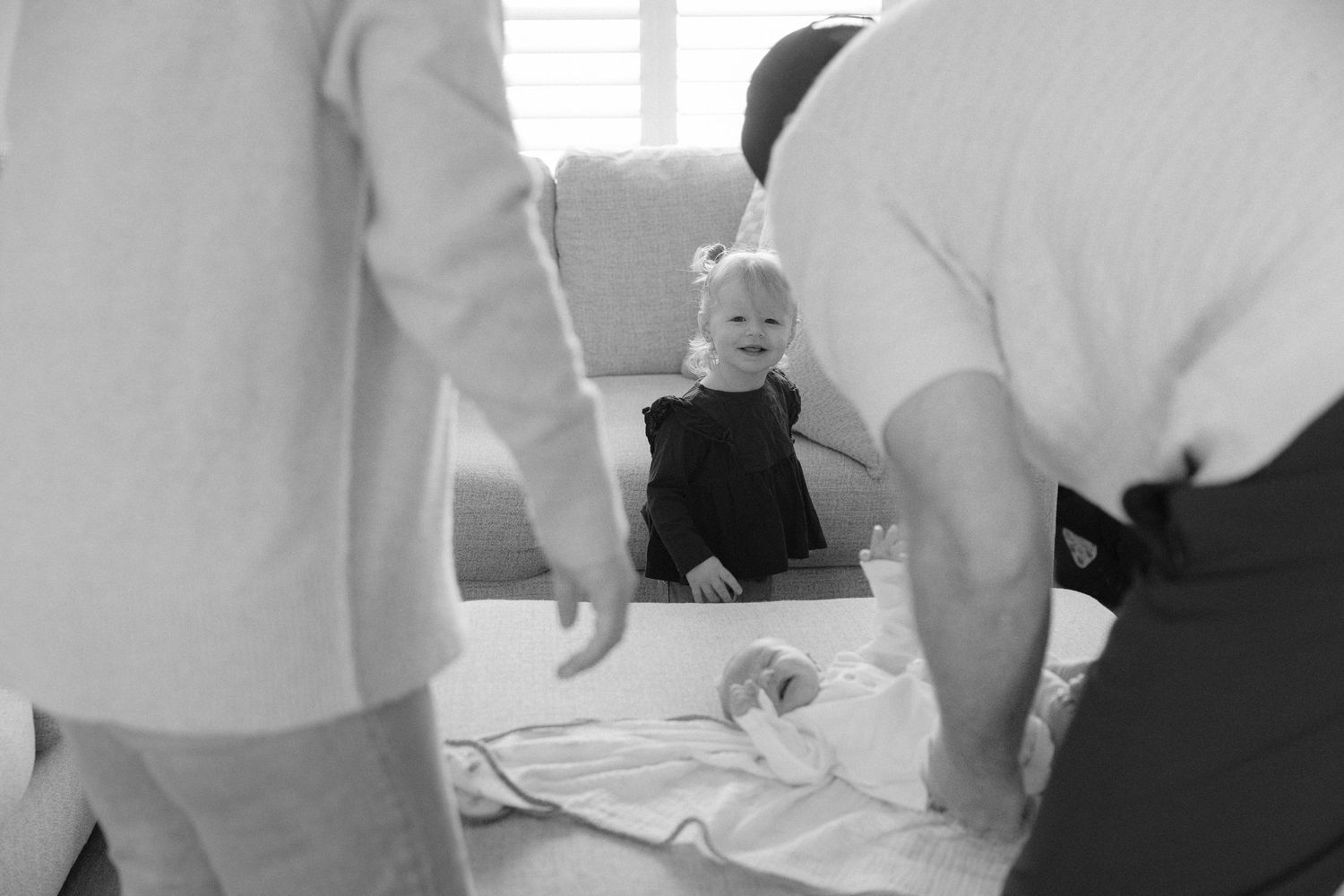 Black and white photo of a smiling toddler watching parents fold a white blanket on the floor.
