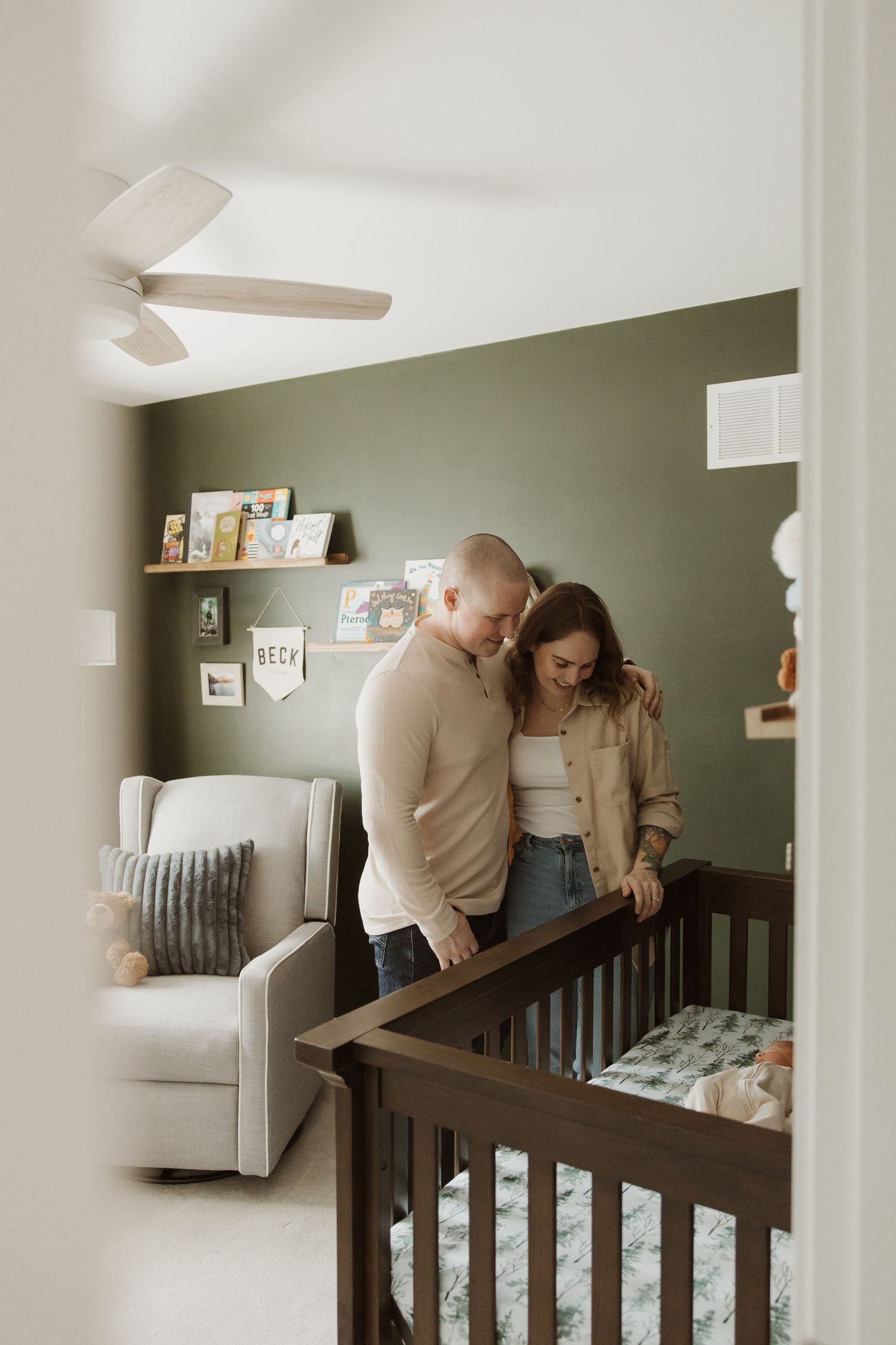 Couple standing together over a dark wood crib in a green nursery room viewed through a doorway.