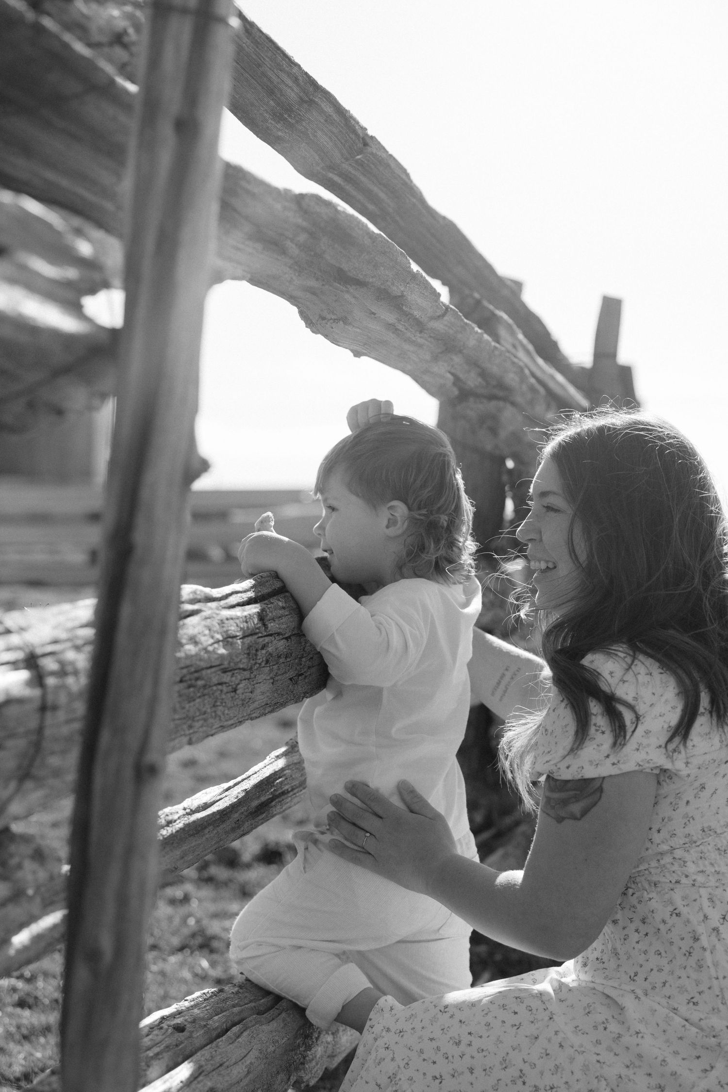 Black and white photo of a smiling mother holding her toddler on a rustic wooden fence.