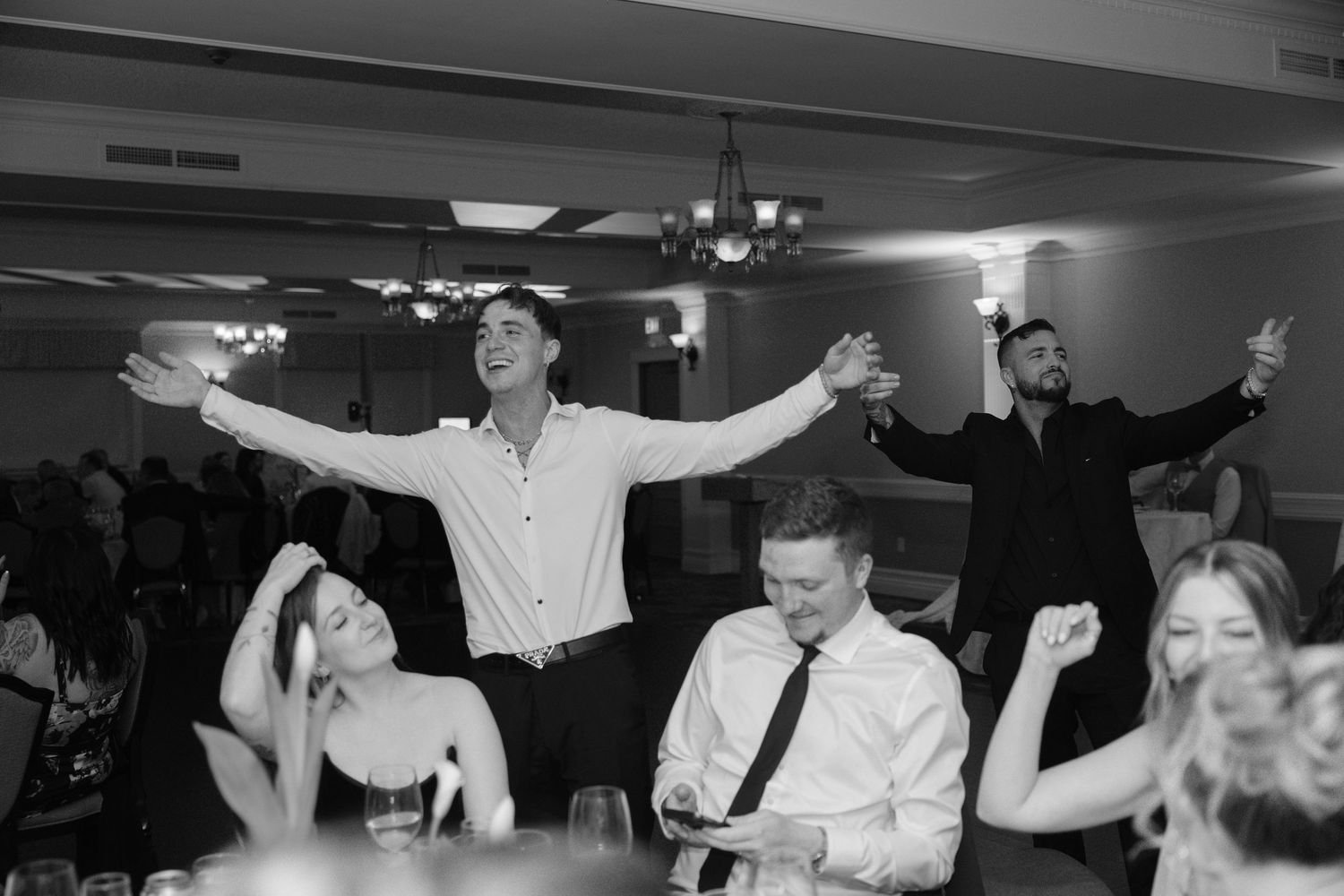 Guests celebrating joyfully at a wedding reception, with a man spreading his arms wide in a black and white photo.