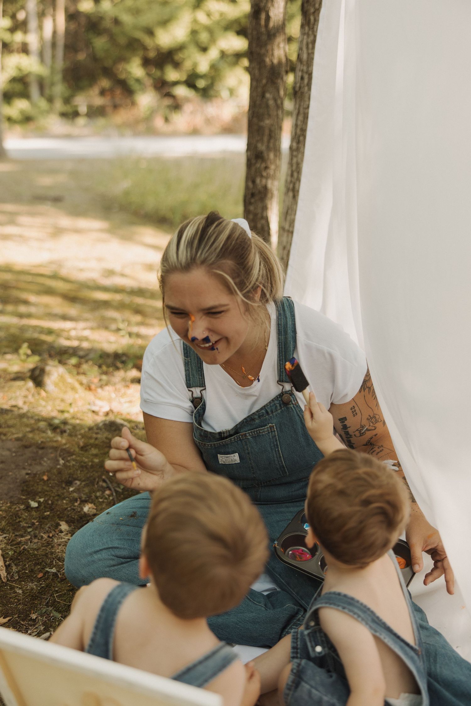 Smiling woman in overalls playing with twin babies under white canopy outdoors.