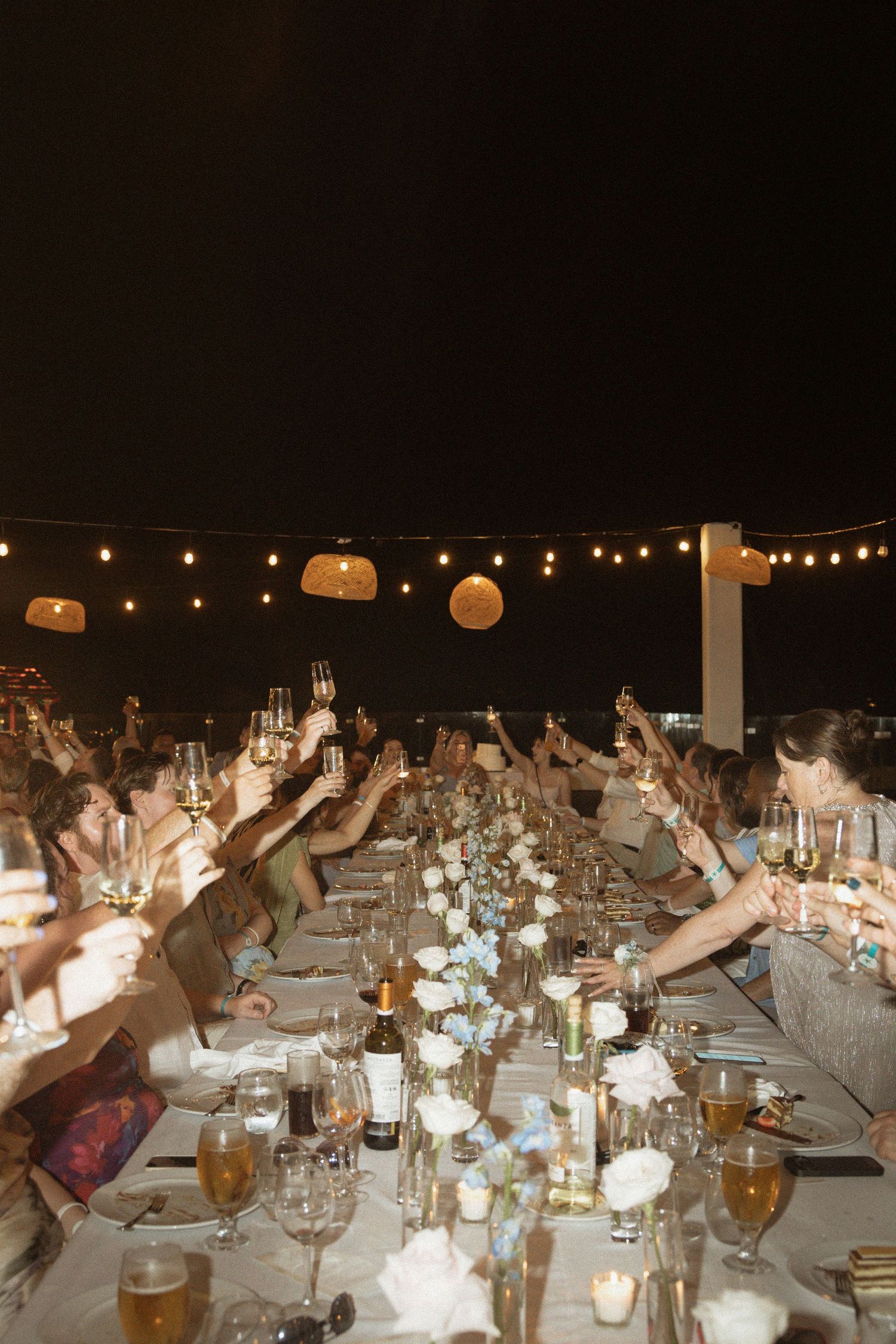 Wedding guests raise champagne glasses in a toast at an elegant outdoor reception dinner under warm string lights.