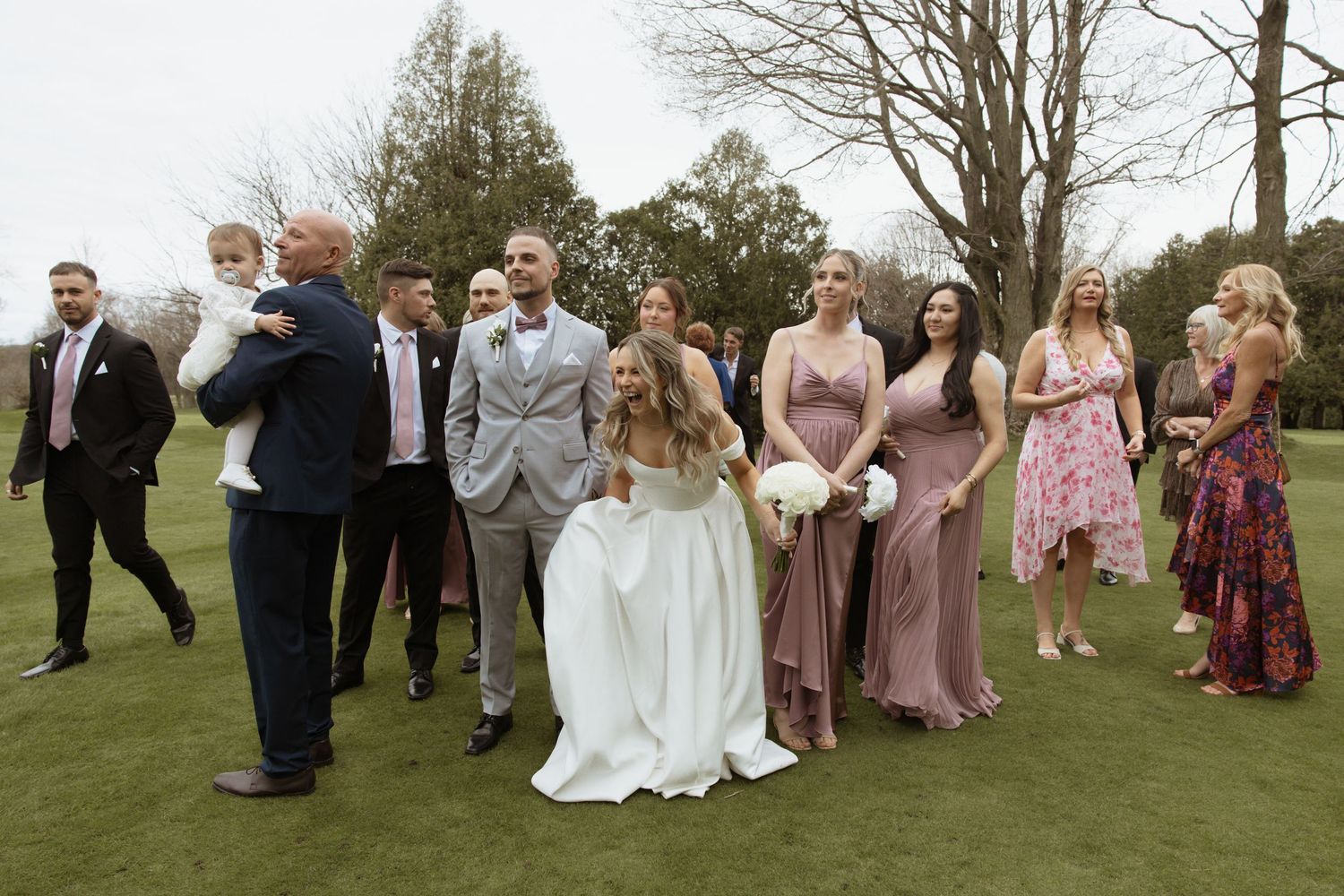 Wedding party group photo on grass with bride, bridesmaids in mauve dresses.