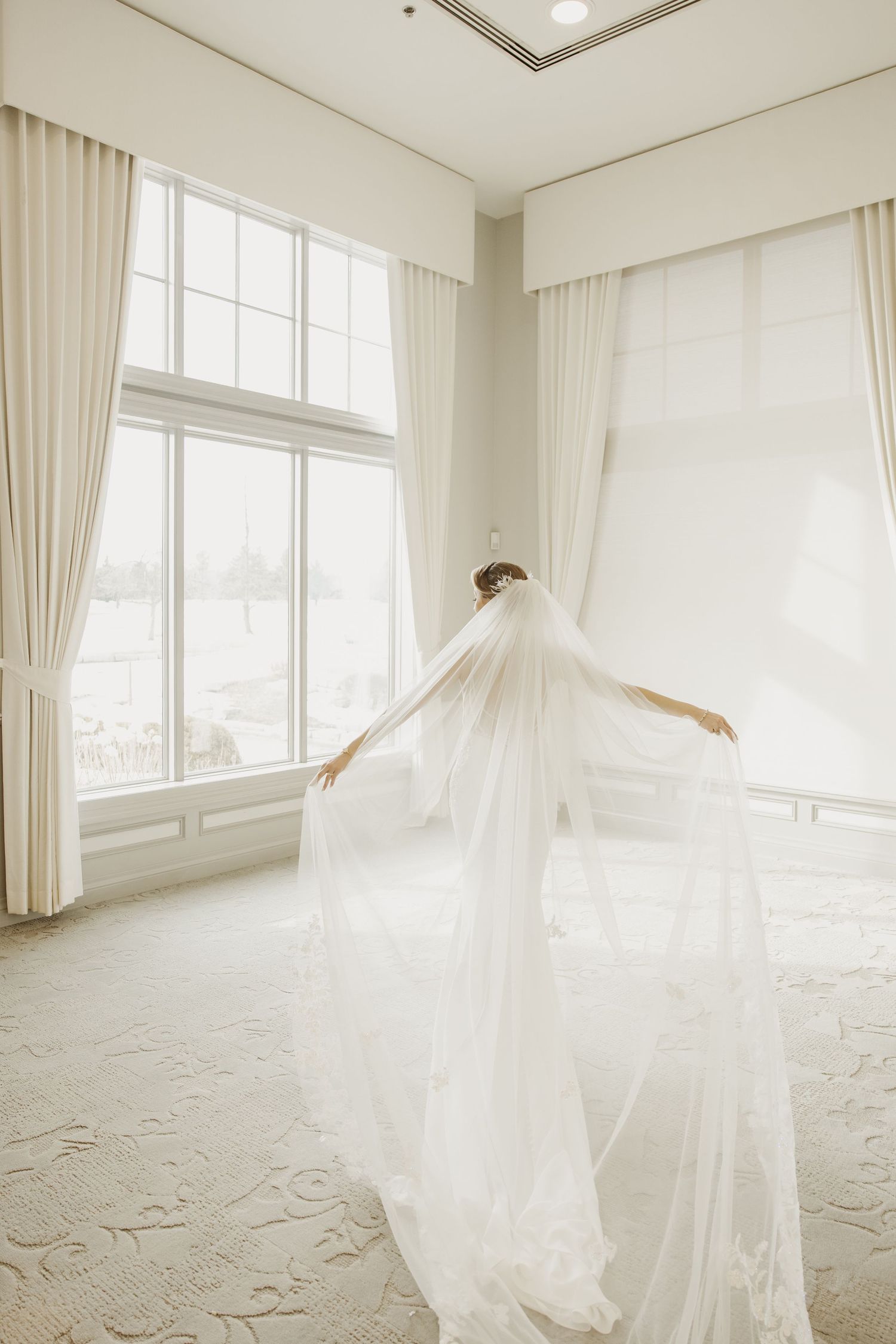 Bride in a flowing white wedding gown and veil posing gracefully in a bright, airy bridal suite.