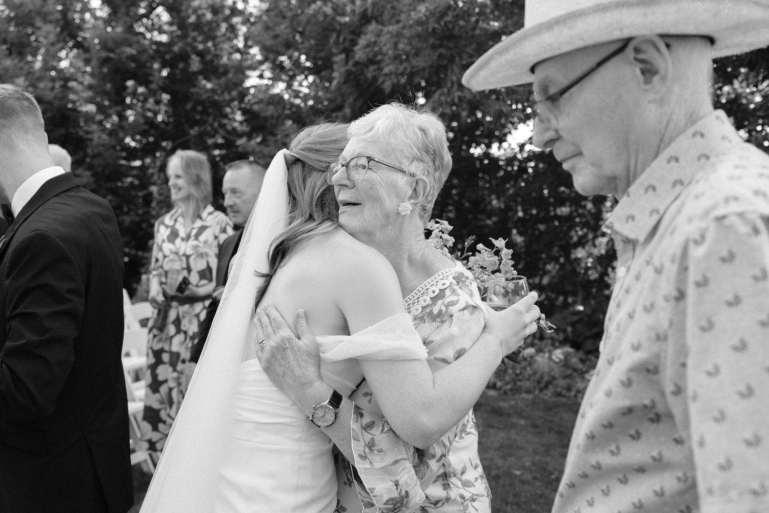 Bride in white gown and veil sharing emotional hug with elderly woman guest at outdoor wedding reception.