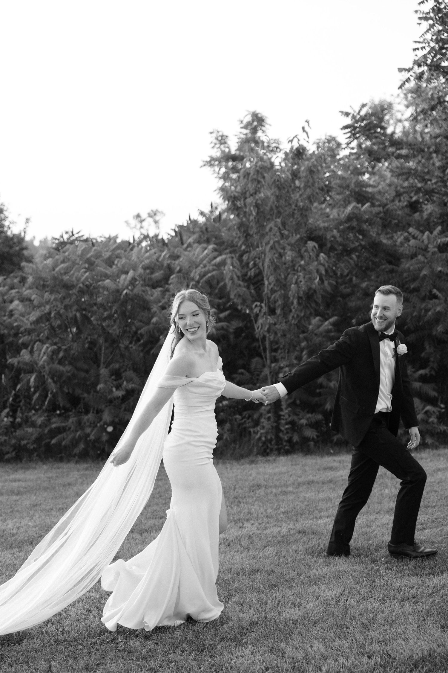 Black and white photo of bride and groom holding hands walking on grass with trees in background.