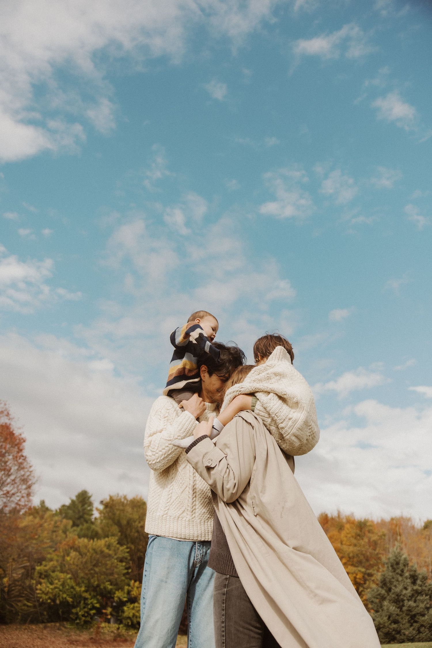 Family embracing outdoors under blue sky with autumn foliage in background.
