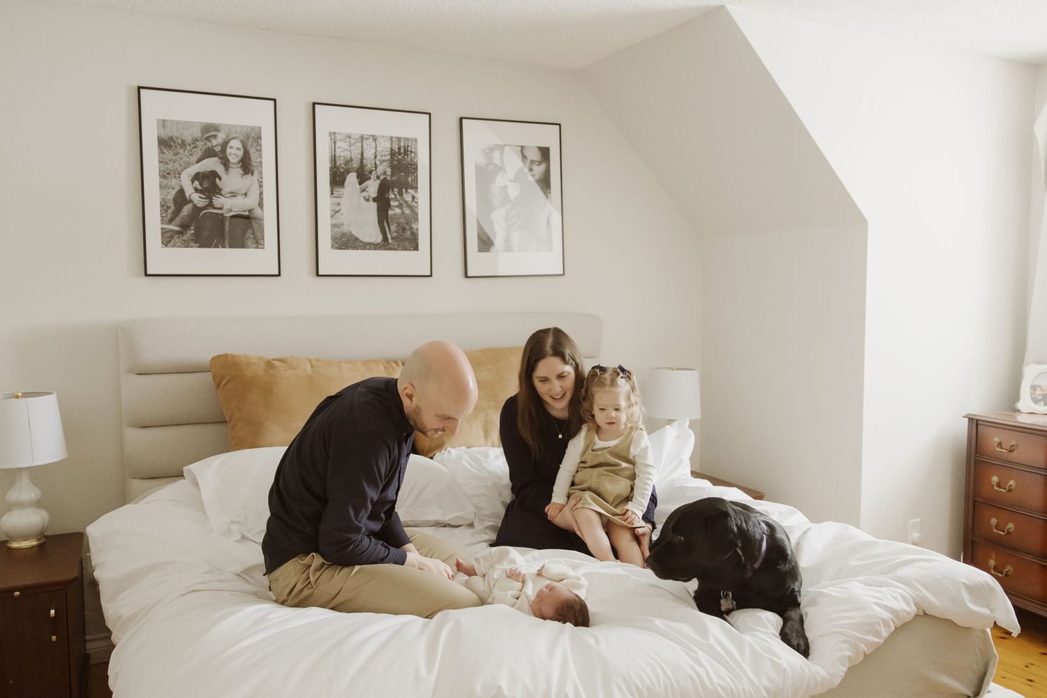 Parents and toddler gathered around their newborn baby on a white bed with a black dog nearby.