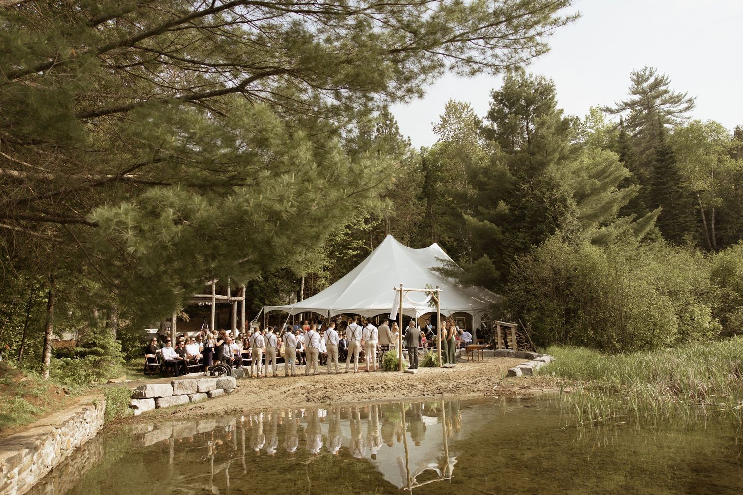 Rustic outdoor wedding reception tent beside a reflective pond surrounded by pine trees.