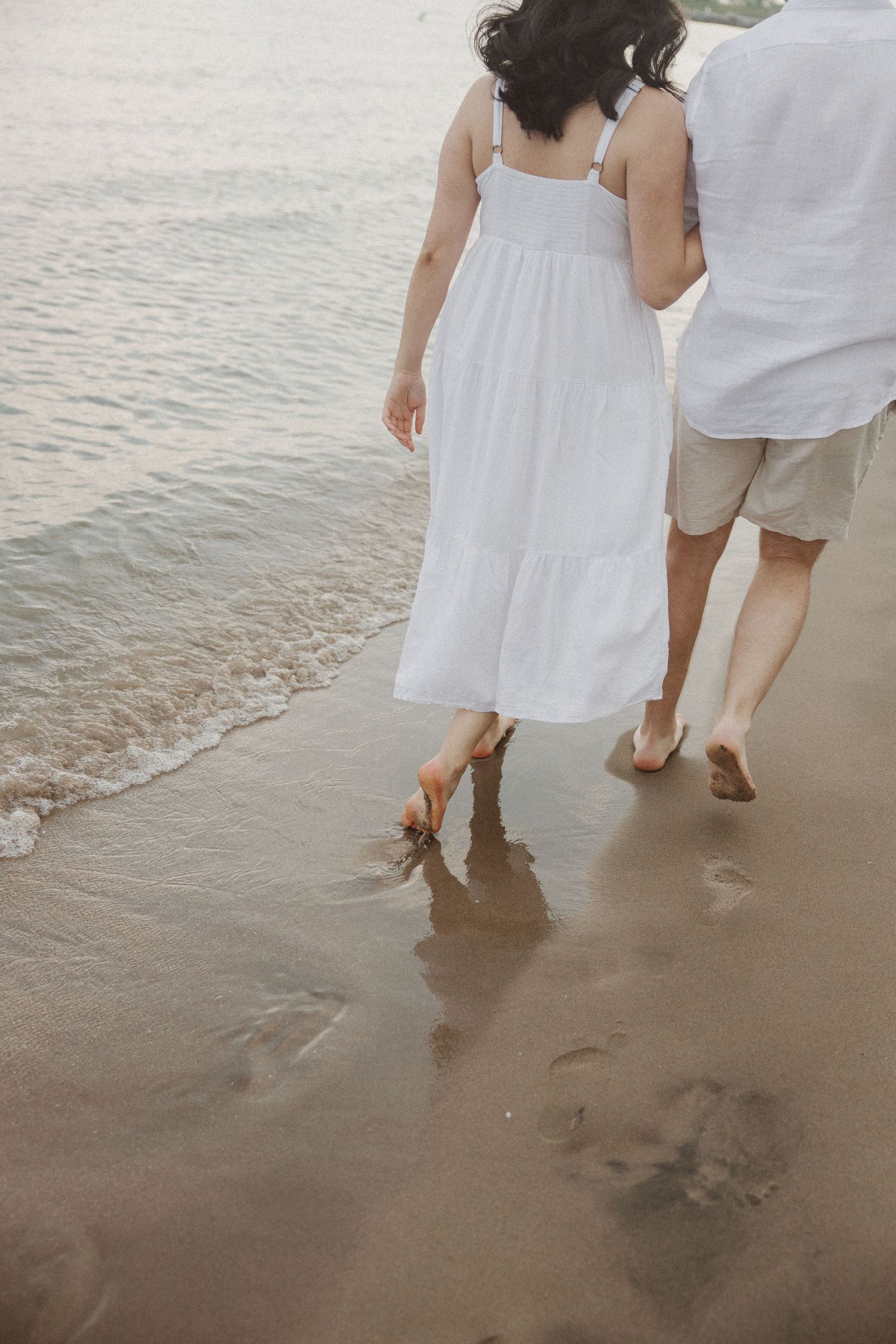 Couple walking barefoot along shoreline, woman in white dress leaving footprints in sand.