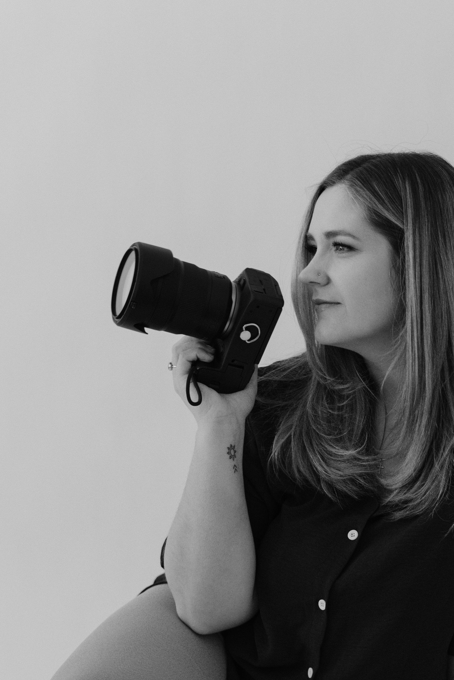 Black and white photo of woman holding DSLR camera up to shoot, small tattoo visible on wrist.