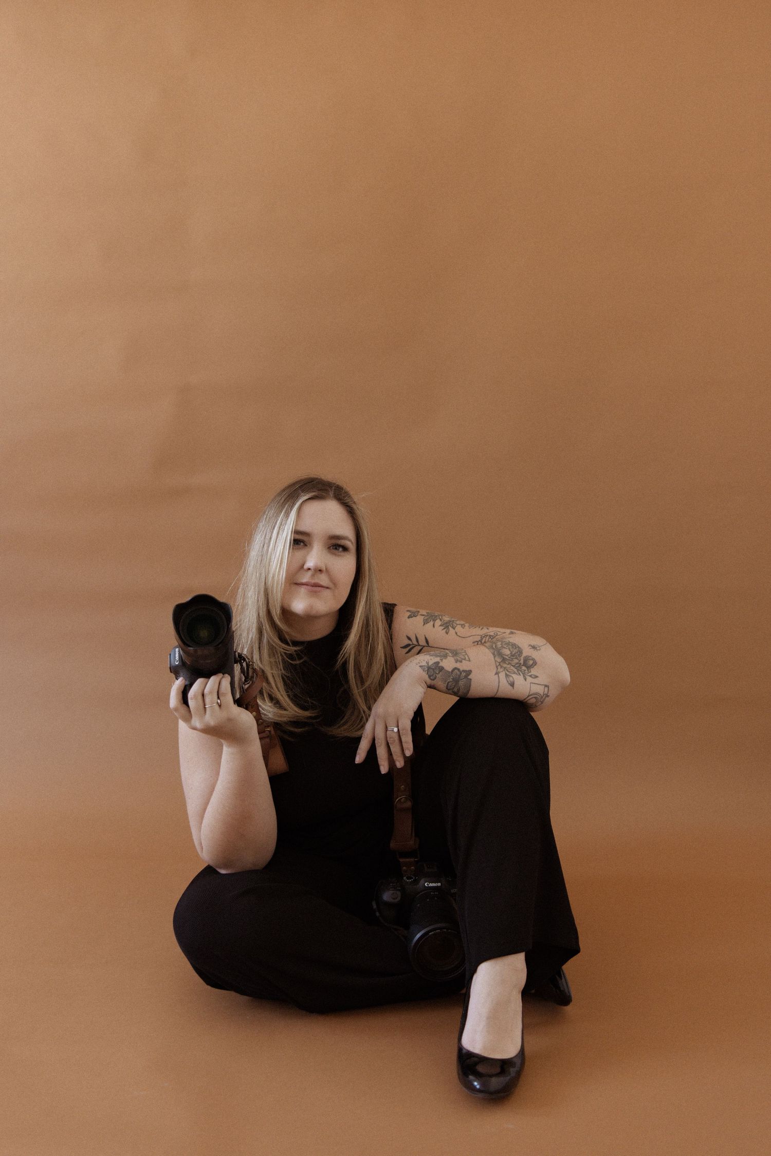 Female photographer sitting on brown background in black outfit holding camera with tattooed arm.