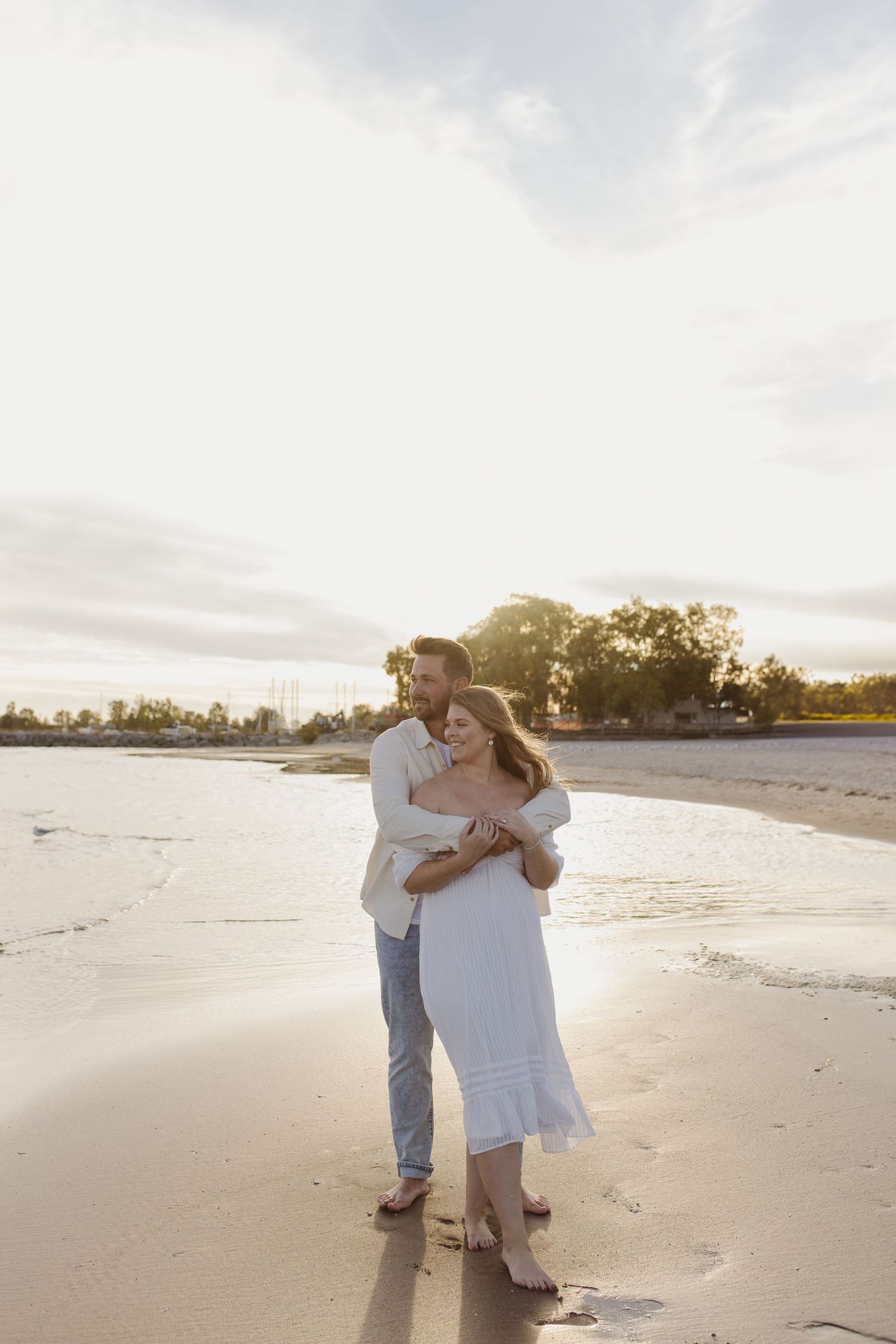 Couple sharing a romantic embrace on a sunlit sandy beach with gentle waves nearby.