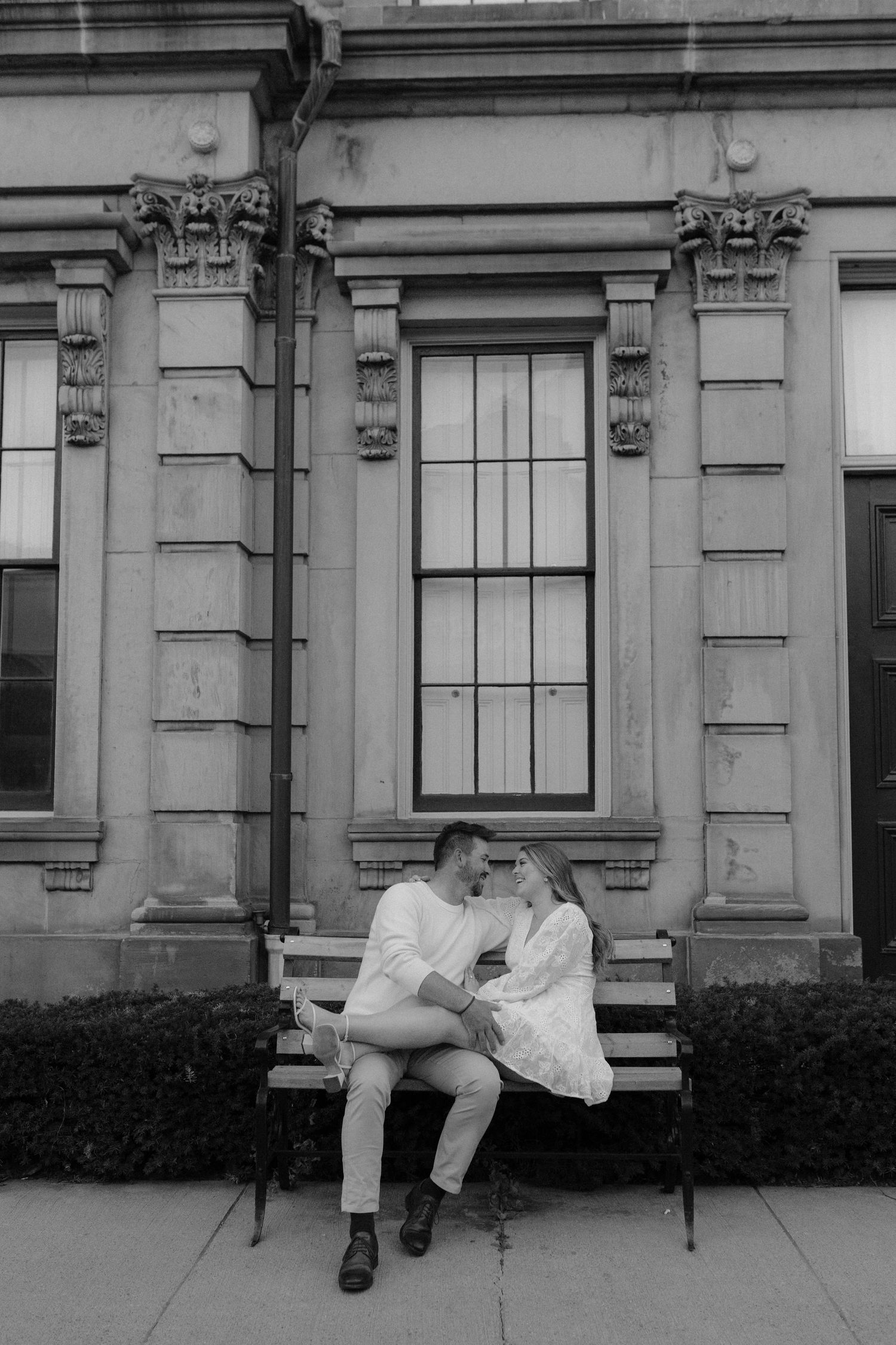 Black and white photo of couple sitting close together on bench outside historic stone building.