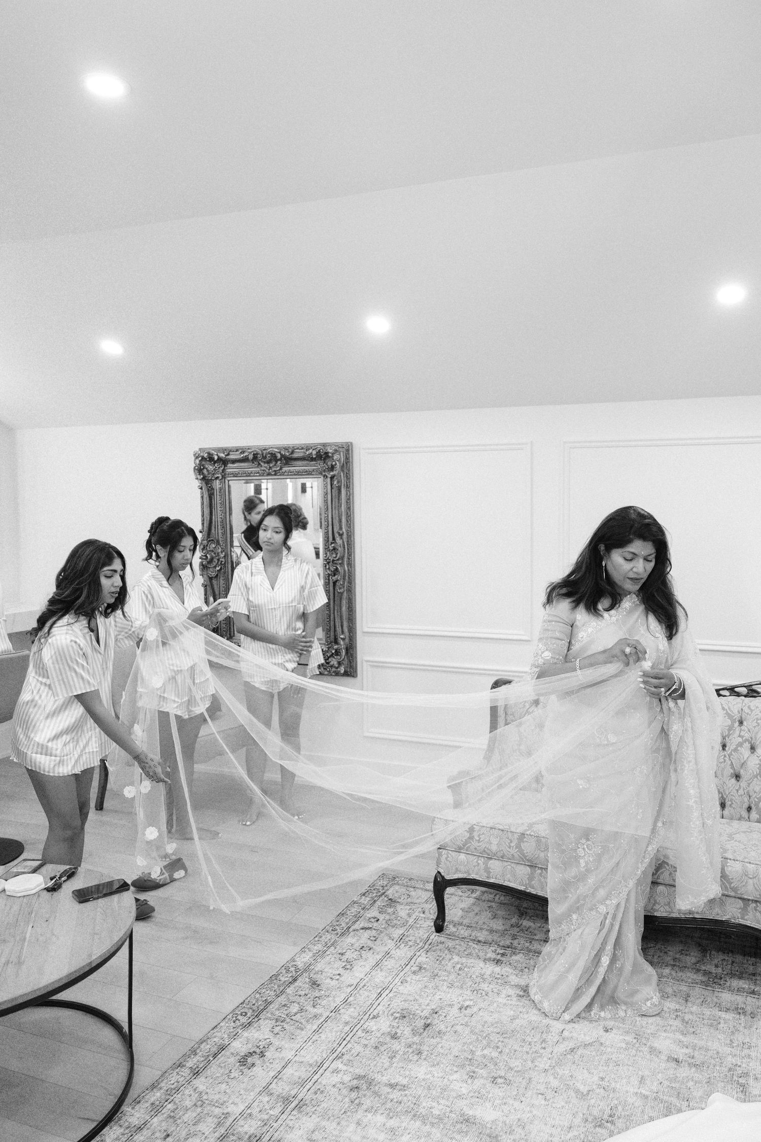 Black and white photo of bridesmaids helping the bride with her long veil in a bright white dressing room.