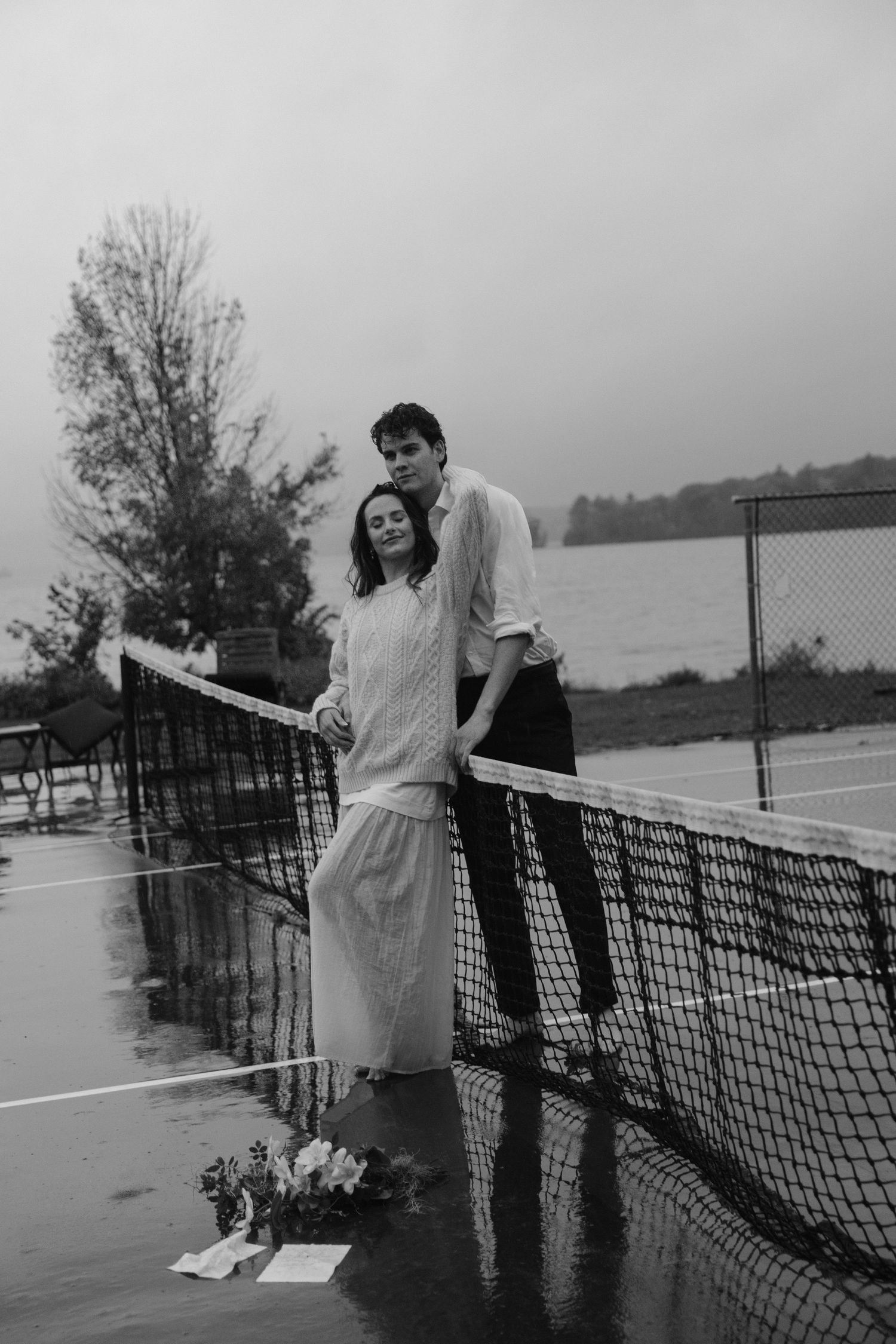 Black and white romantic couple embracing at tennis net in the rain.