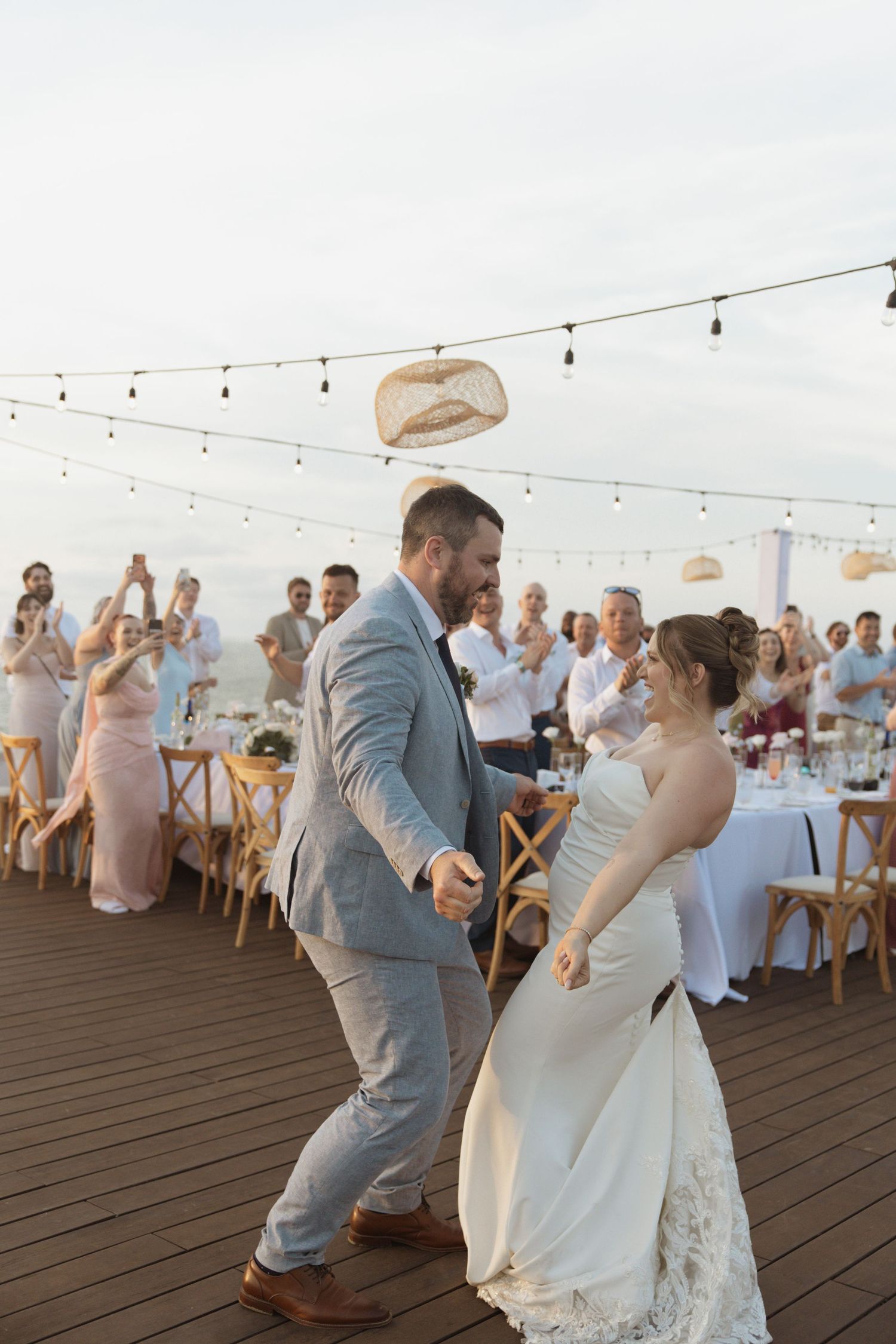 Bride and groom share first dance on outdoor deck at sunset wedding reception with string lights and guests.