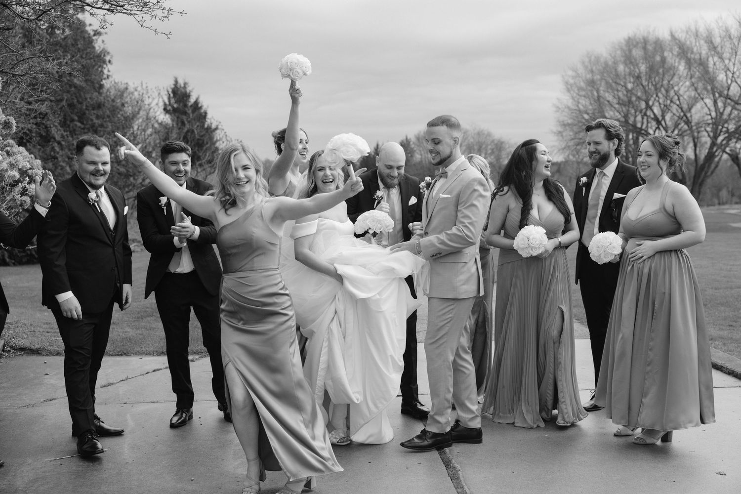 Black and white wedding party photo with bride being lifted joyfully outdoors.