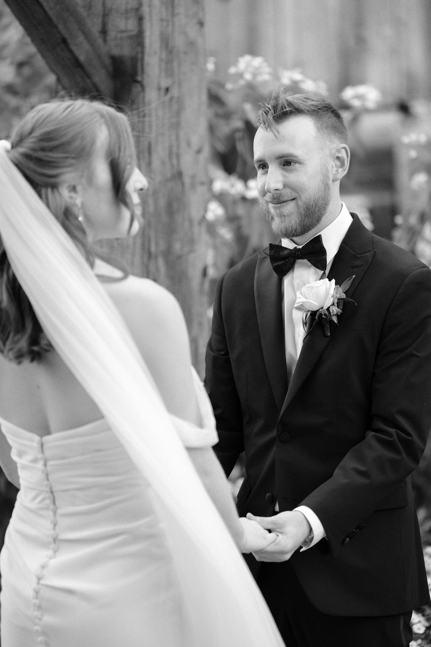 Black and white photo of a groom smiling at his bride during their outdoor wedding ceremony.
