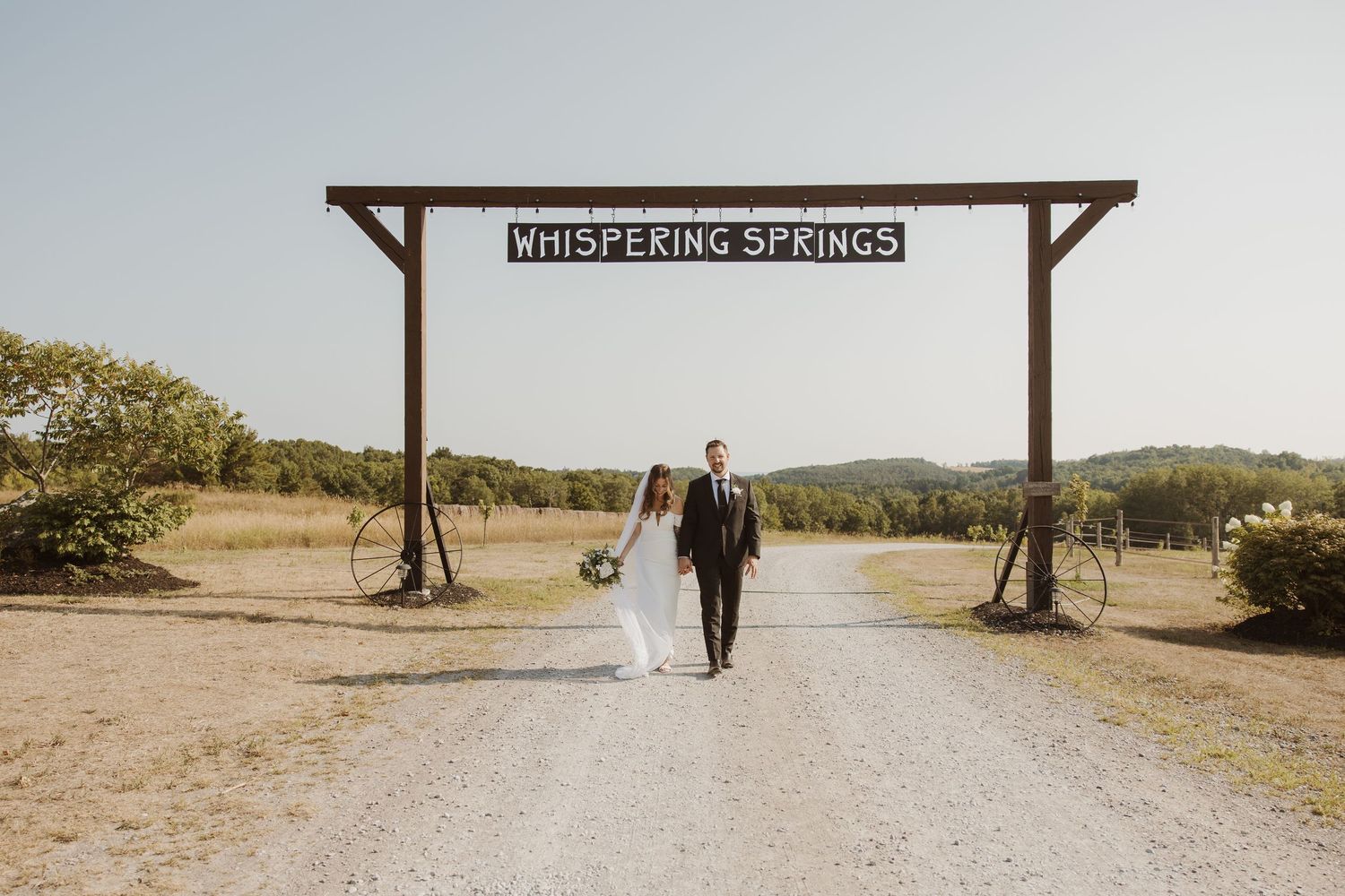 Bride and groom walking hand in hand under a rustic wooden Whispering Springs wedding venue entrance sign.