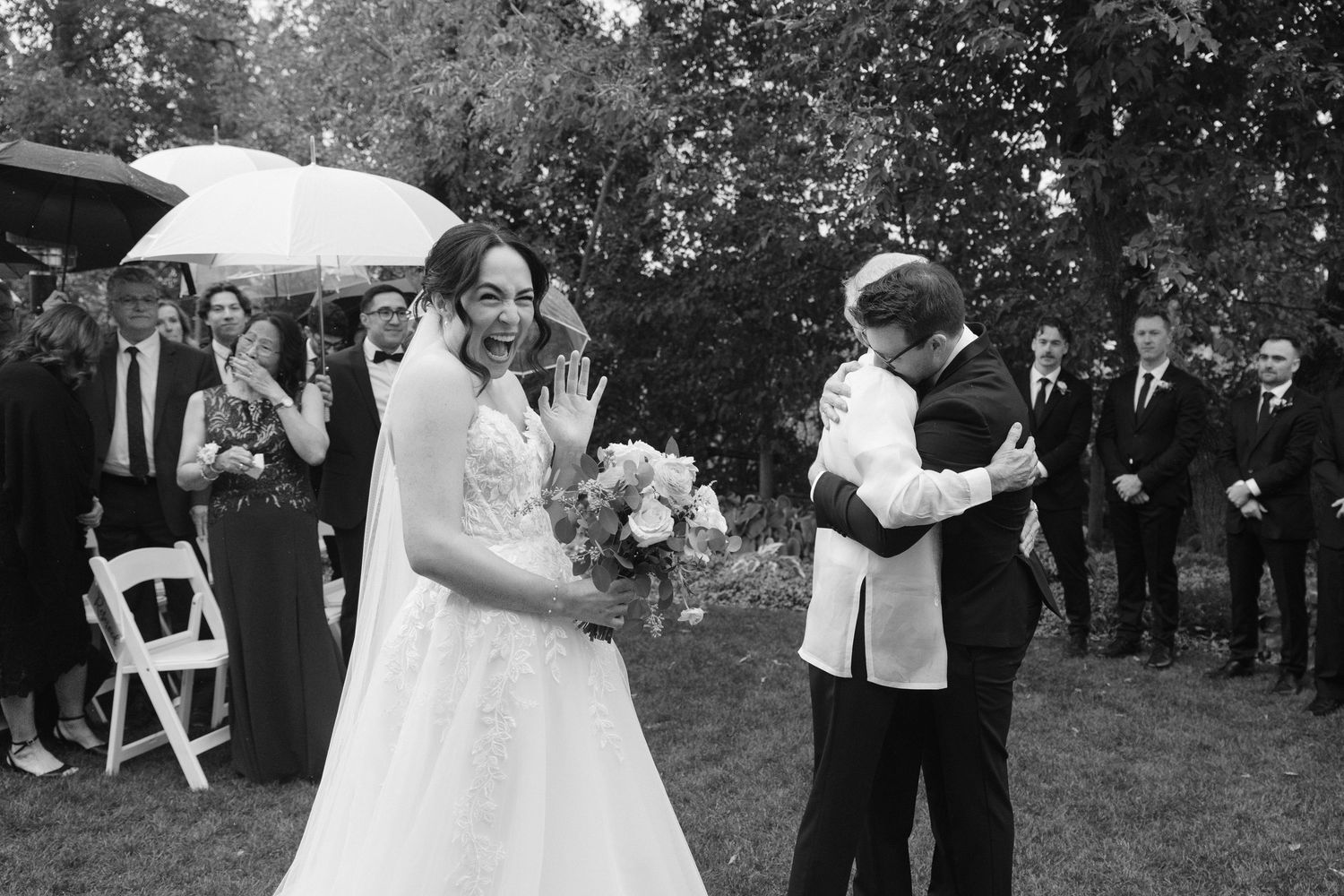 Black and white photo of joyful newlyweds just married, bride holding bouquet under umbrella outdoors.