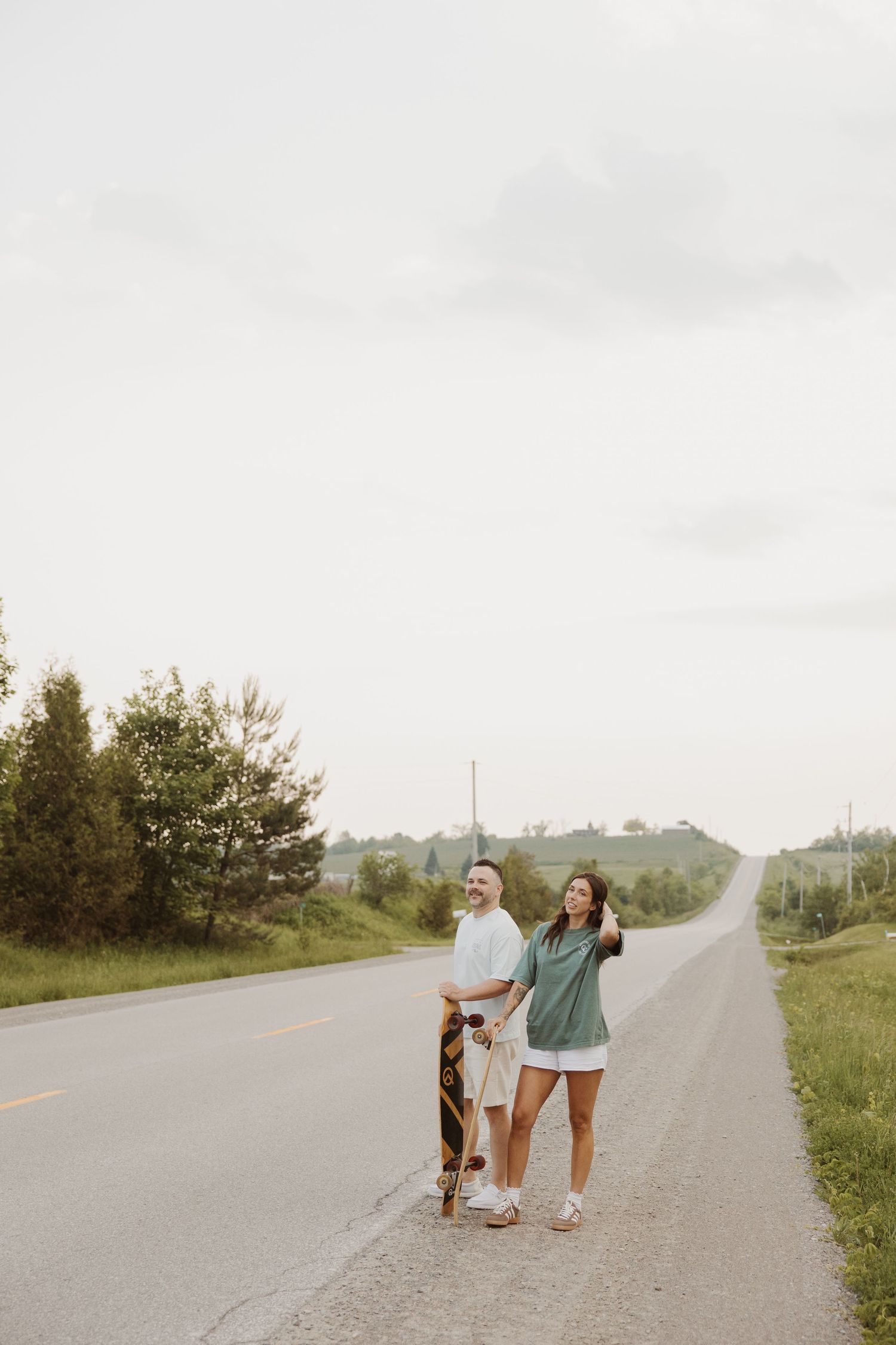 Engaged couple standing on rural road holding a longboard skateboard.