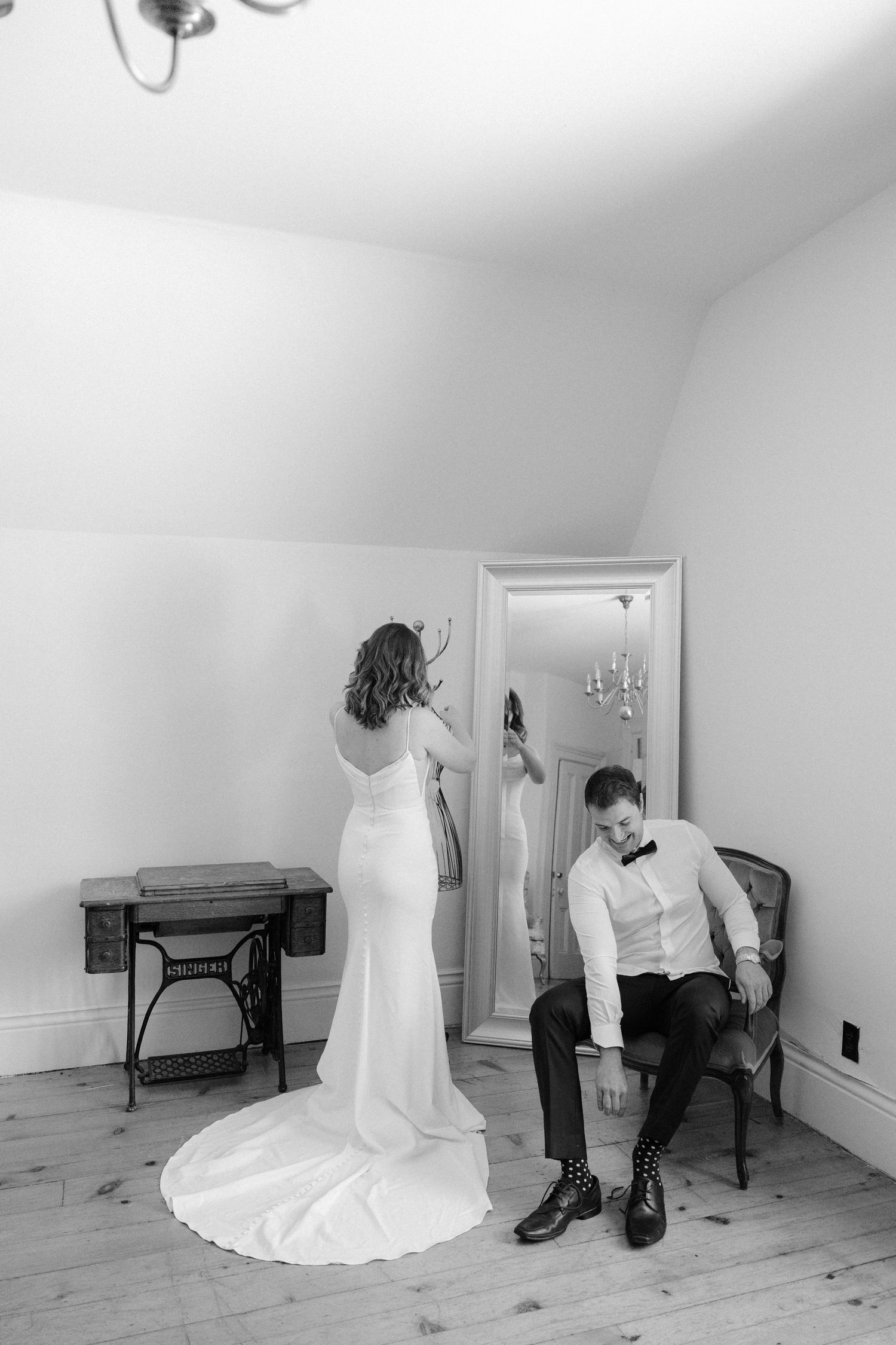 Bride and groom getting ready in black and white, she adjusts hair as he ties shoes.