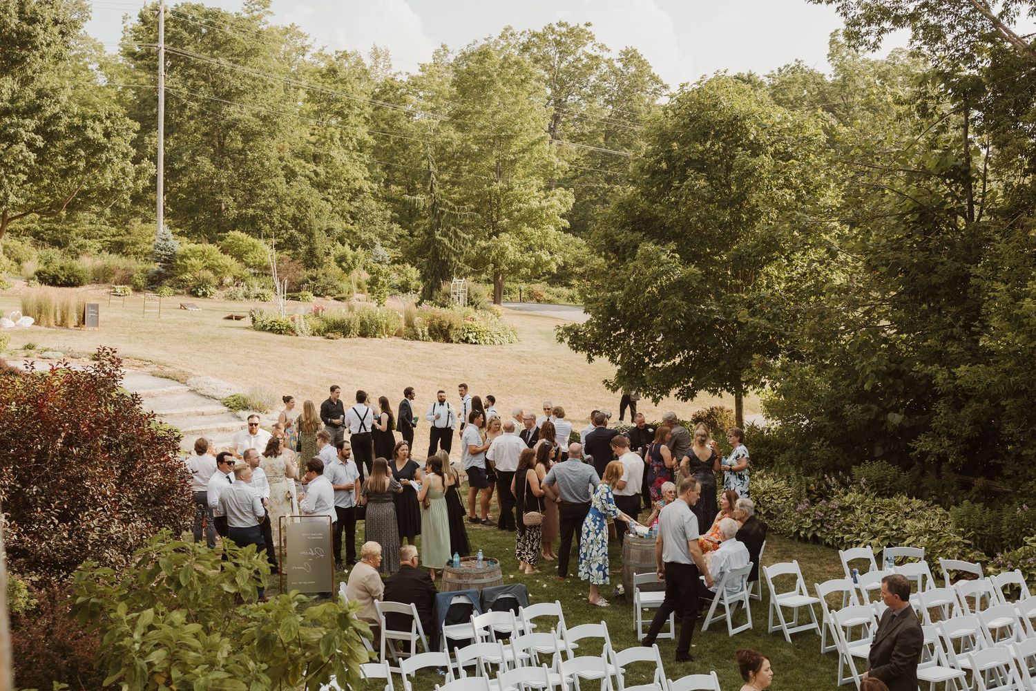 Aerial view of wedding guests mingling outdoors among white chairs and lush green trees.