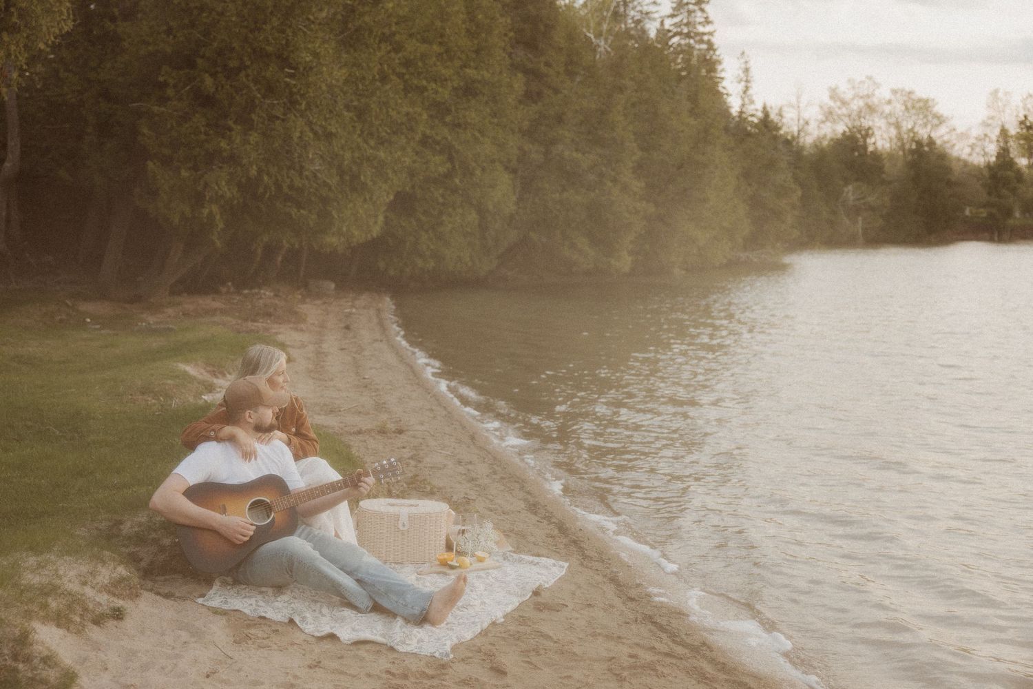Romantic couple sitting on a sandy beach blanket by the water, man playing guitar at golden hour.