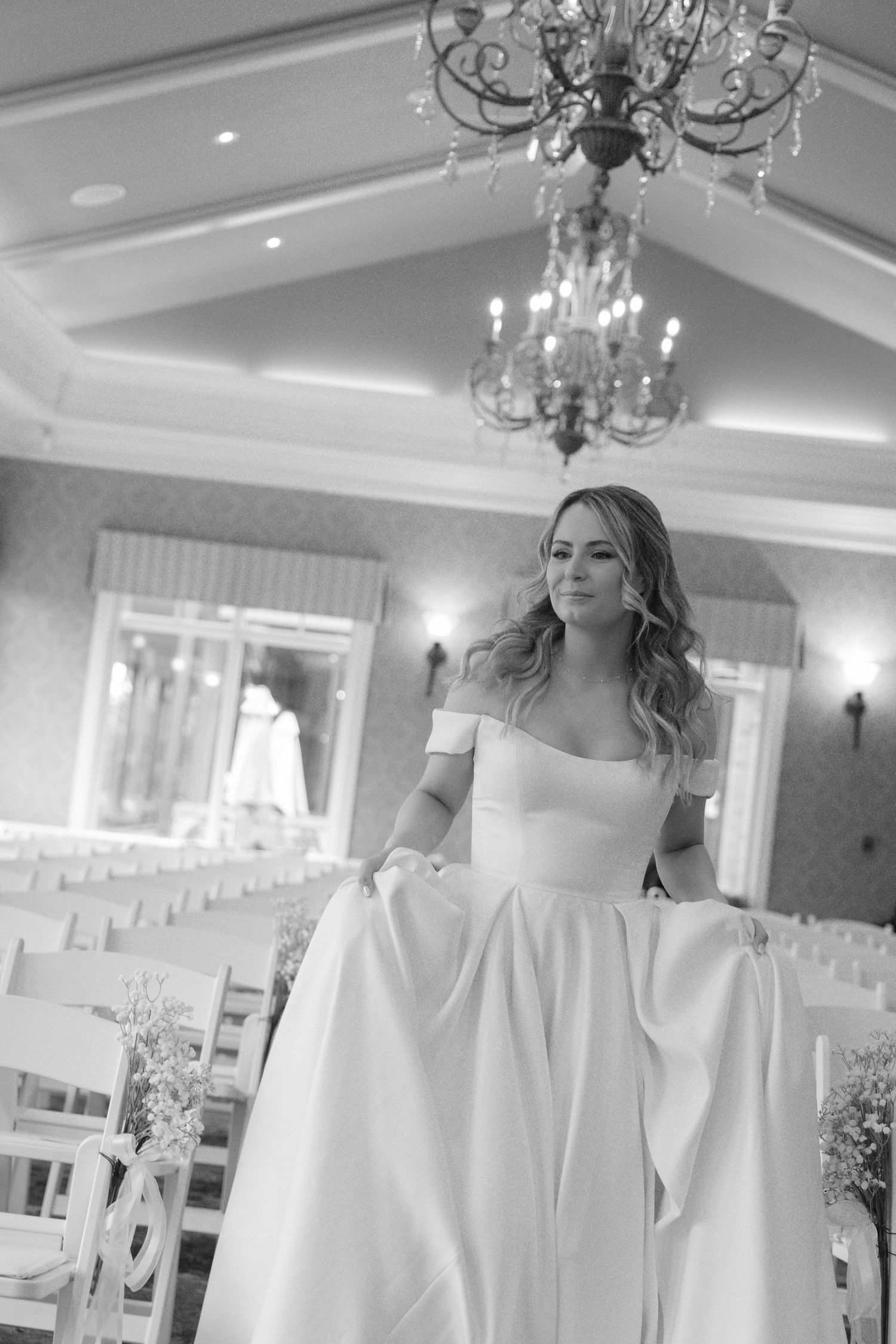 Black and white photo of a bride in an off-shoulder ball gown holding her skirt in an elegant chandelier-lit wedding venue.