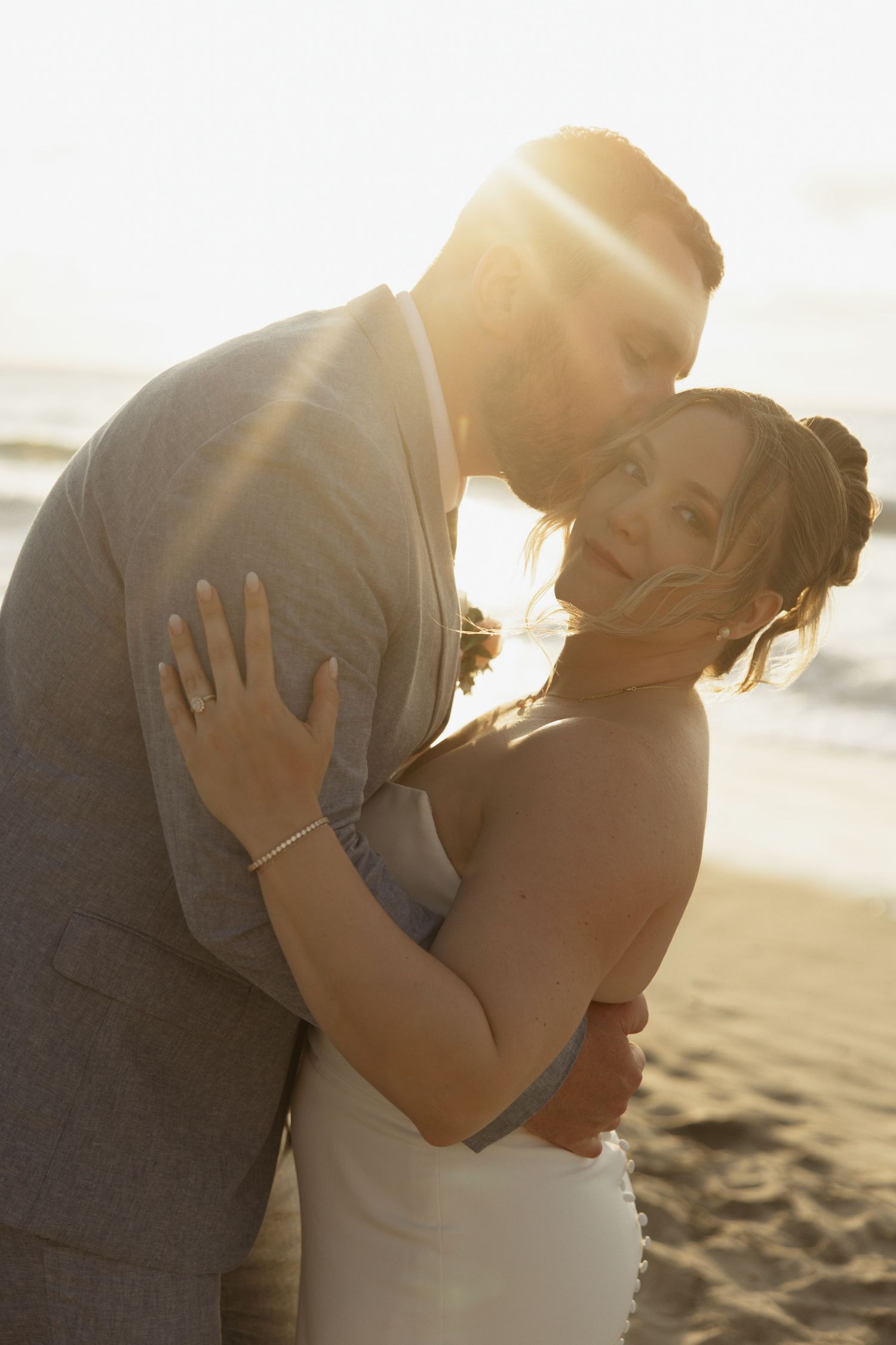 Bride and groom embrace romantically on beach at golden sunset with warm lens flare.