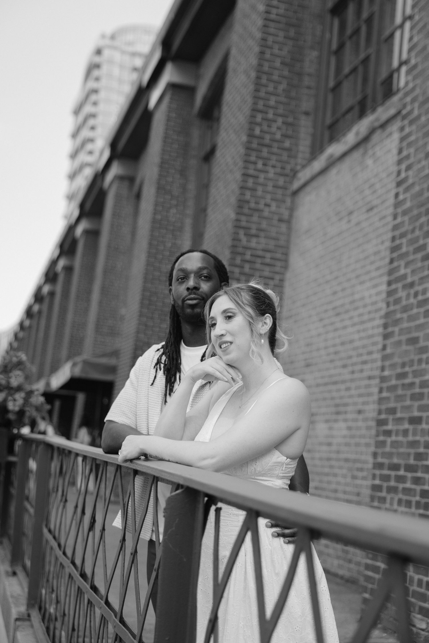 Black and white portrait of interracial couple leaning on a railing outside a brick building in the city.