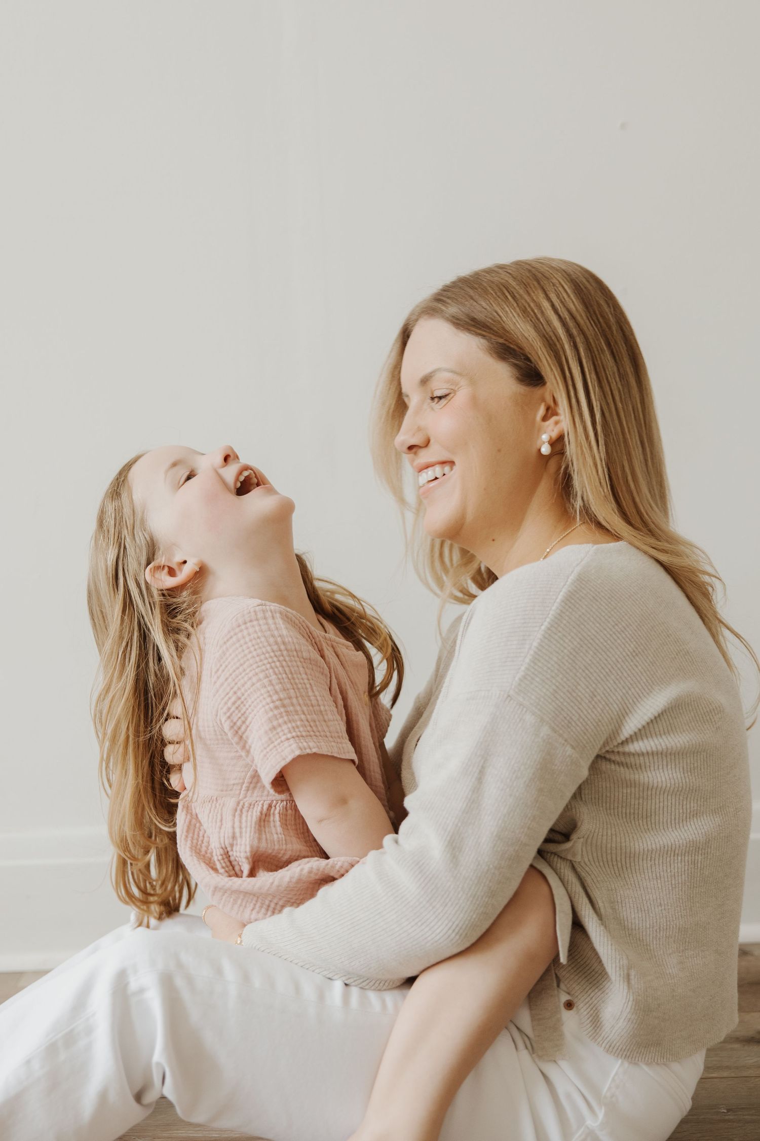 Joyful mother and young daughter laughing together while sitting against a light neutral background.