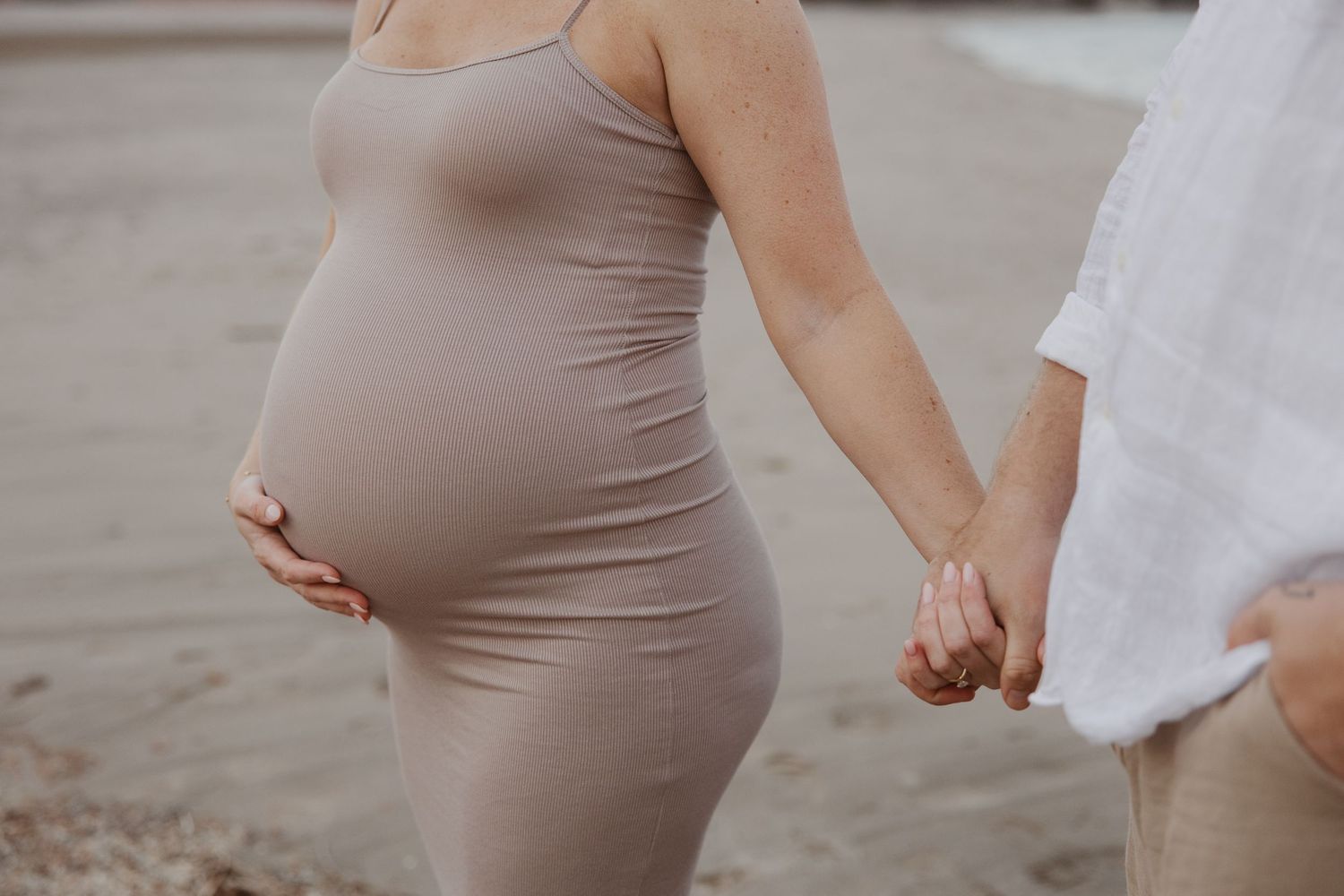 Close-up of a pregnant woman in a beige dress holding hands with her partner on a sandy beach.