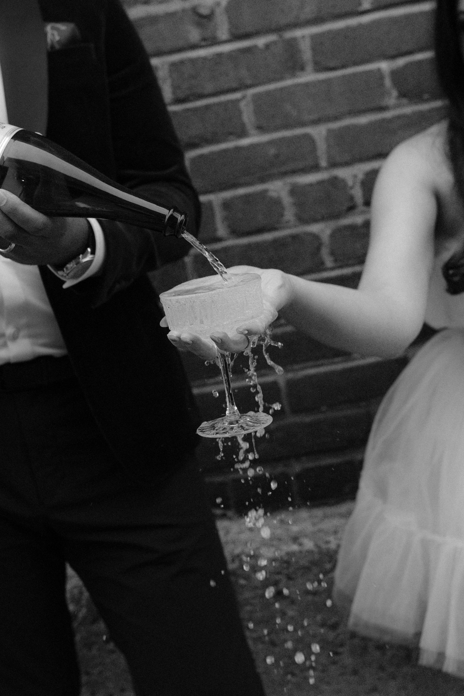 Champagne overflows from stacked glasses as groom pours bottle at wedding in black and white.