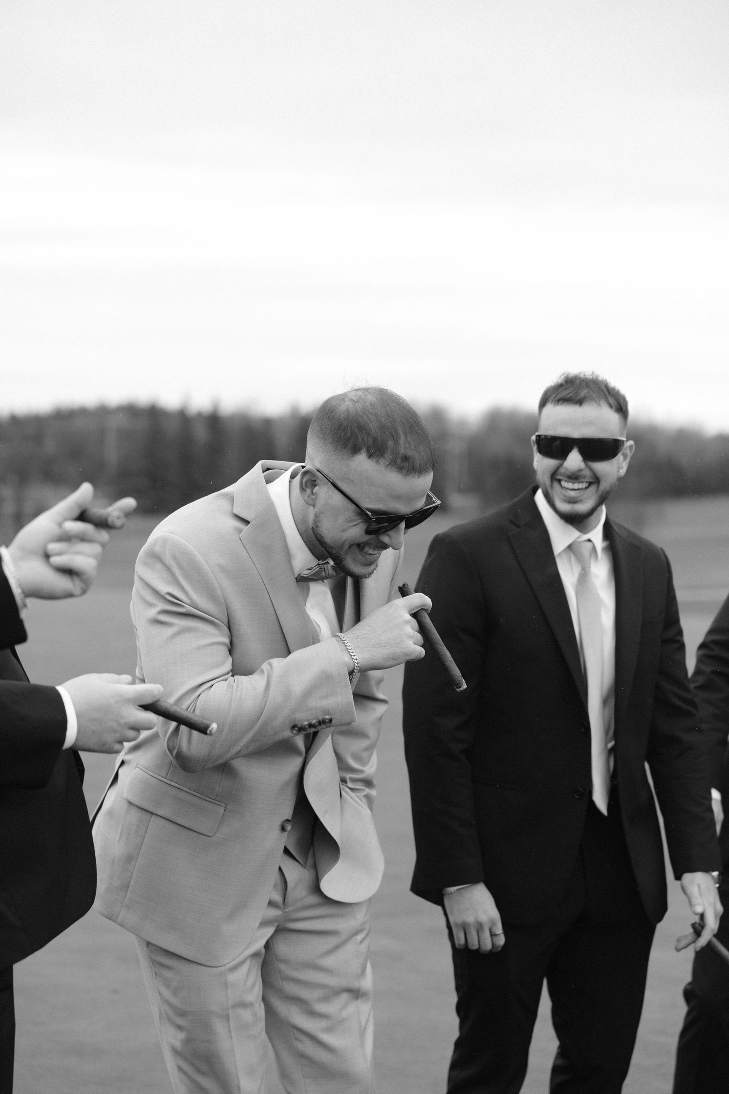 Black and white photo of groomsmen in suits and sunglasses laughing and smoking cigars outdoors.