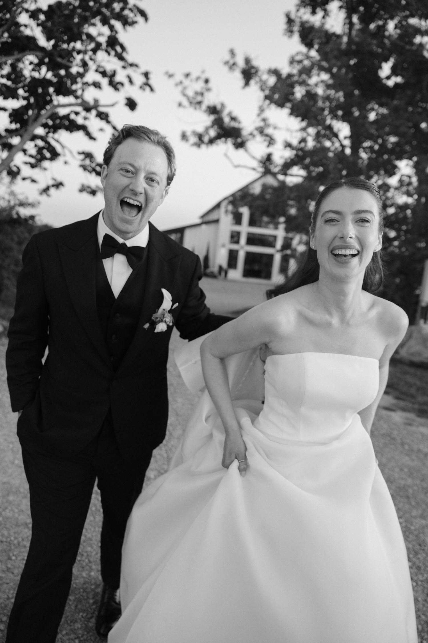 Black and white photo of a laughing bride and groom in formal attire celebrating outside a white building.