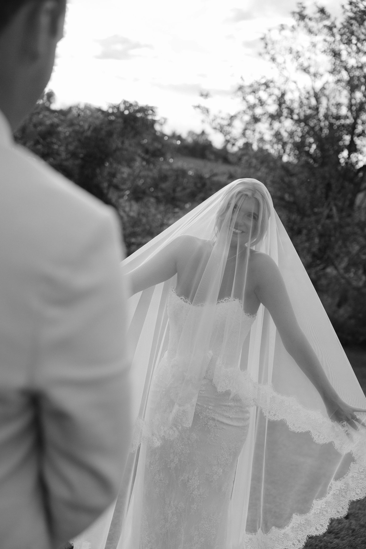 Black and white photo of a smiling bride in a lace wedding dress and long veil, facing her groom outdoors.
