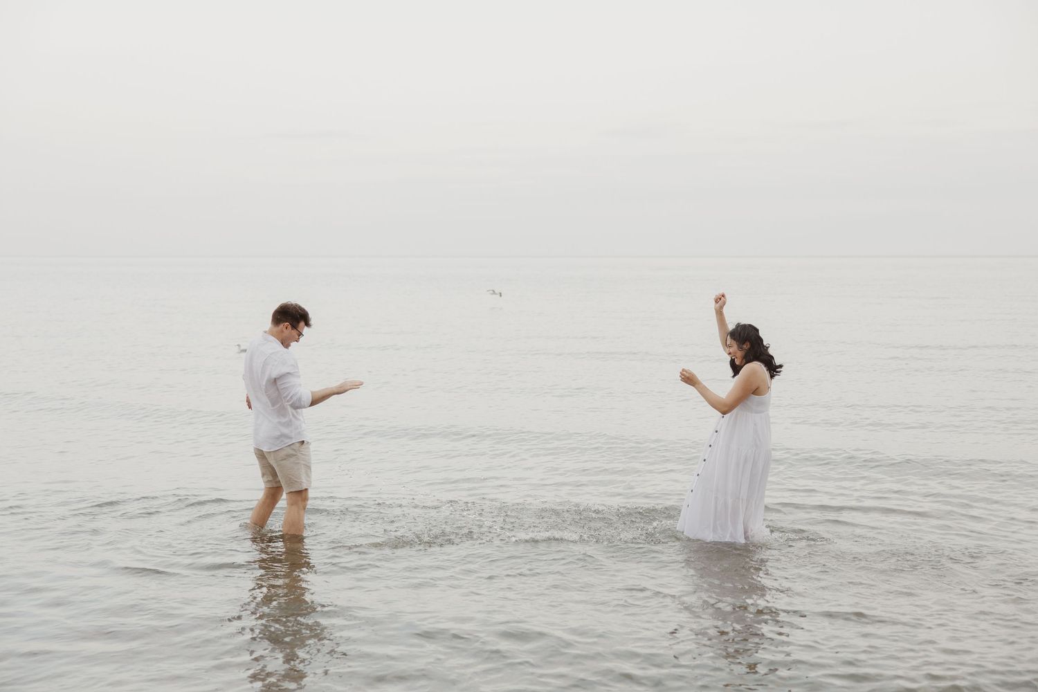 Playful couple splashing and dancing together in calm shallow ocean water on an overcast day.