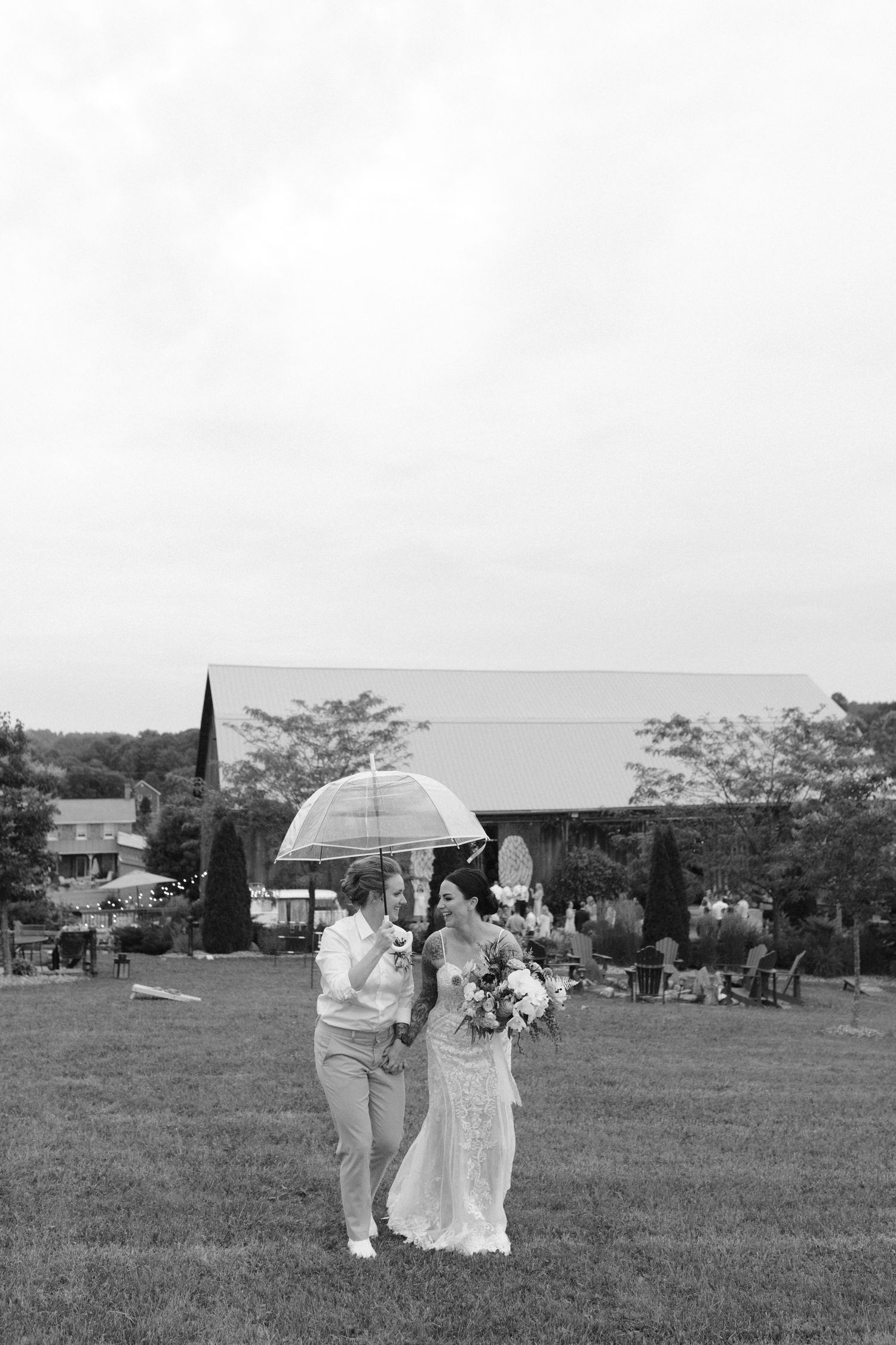 Black and white photo of same-sex couple sharing an umbrella on a rainy wedding day on a farm.