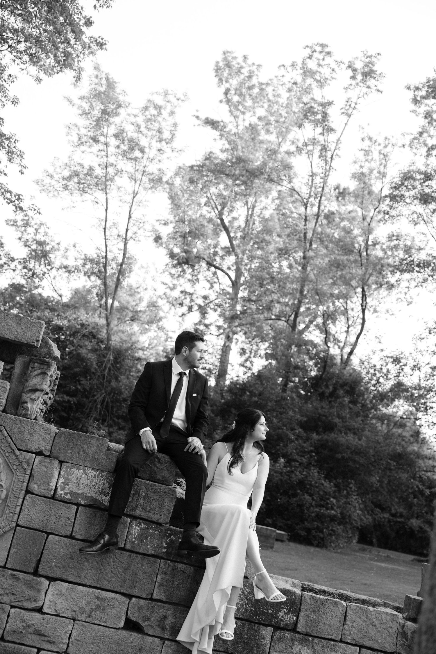 Black and white photo of bride and groom sitting on stone wall surrounded by tall trees in park.