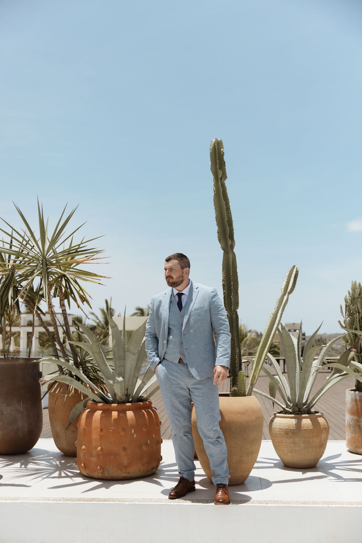 Dapper groom in light blue three-piece suit posing among large terracotta pots with cacti under a bright blue sky.