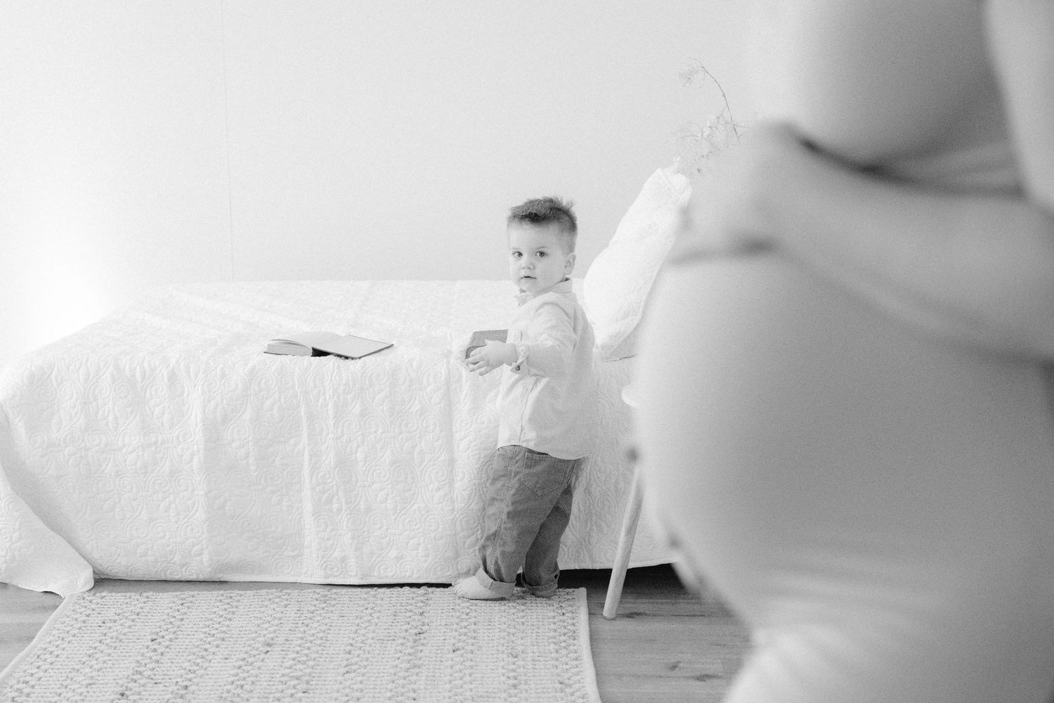 Black and white photo of a toddler standing by a bed near a pregnant woman in a bright room.