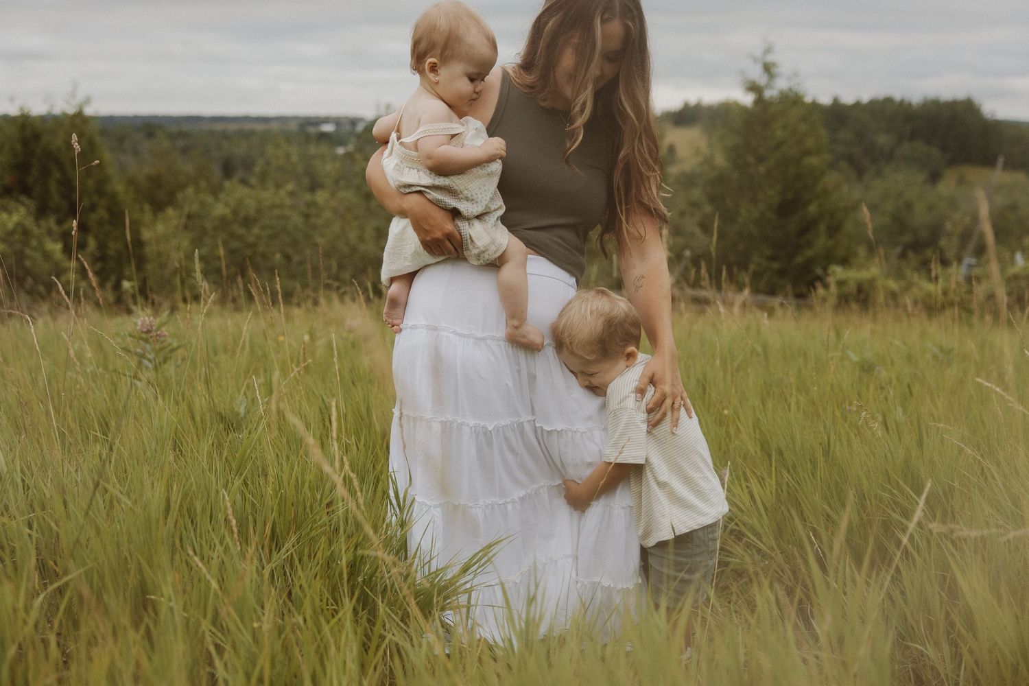 A mother holds her baby while her toddler hugs her in a lush green meadow.